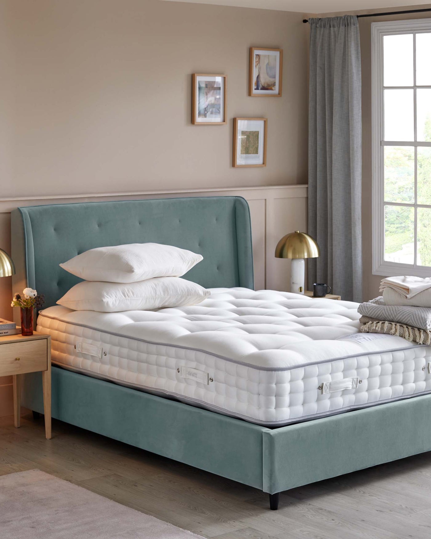 A luxurious plush teal upholstered bed with a button-tufted headboard and a thick white mattress. A light wooden bedside table with a small drawer, topped with a vase of flowers and a gold-coloured lamp, is placed to the left of the bed.