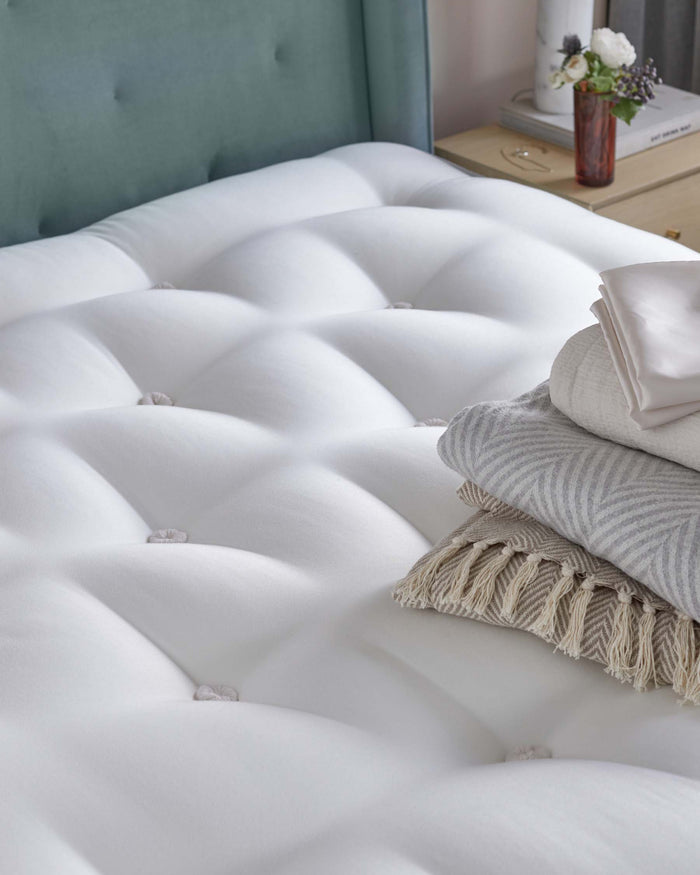 Luxurious white tufted upholstered bench with plush cushioning and small detail buttons, paired with a soft grey and white striped throw blanket and pale pillows on top, beside a pastel green upholstered headboard and a bedside table with a floral arrangement and books.