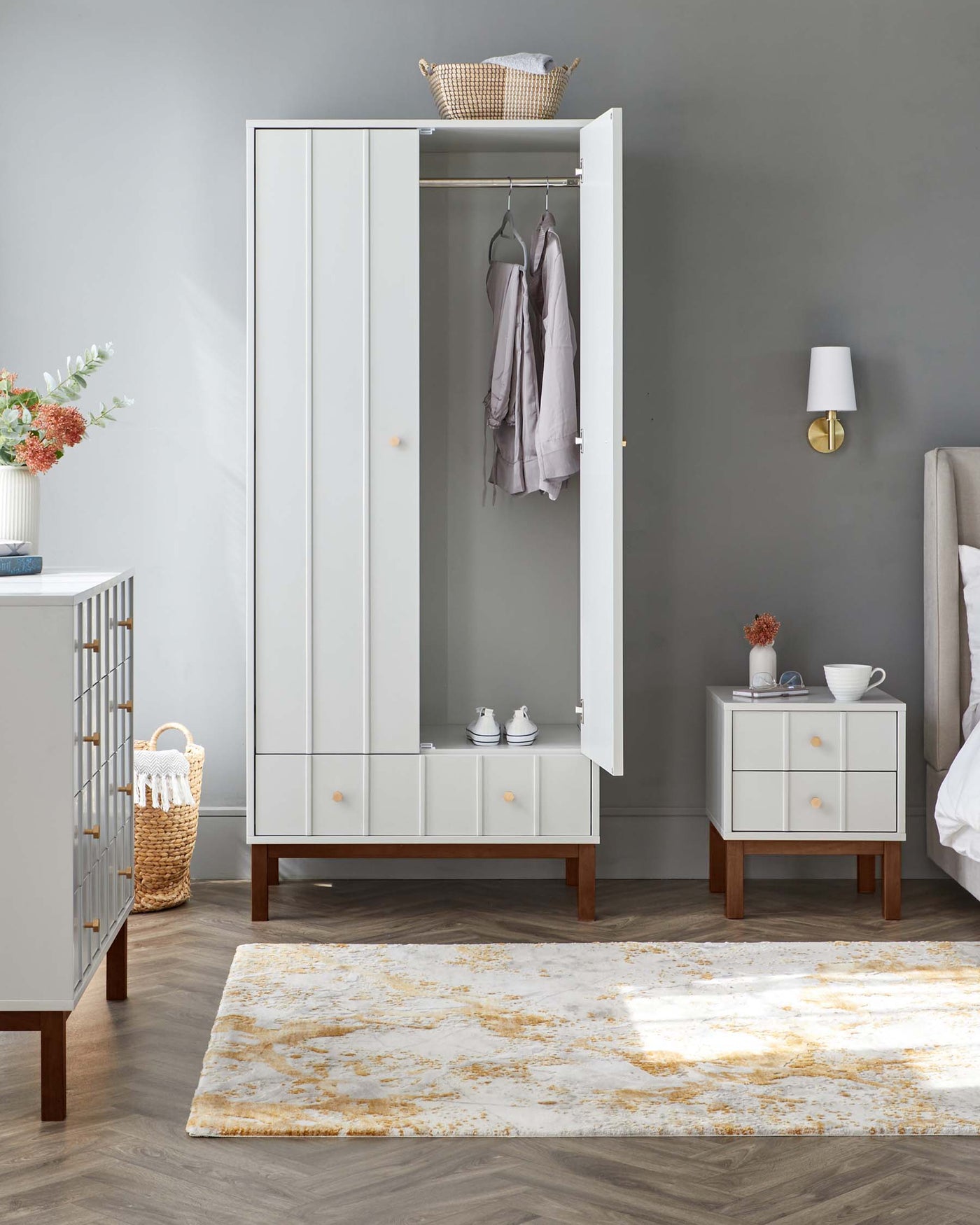 A modern light grey wardrobe with a full-length mirror, wooden base, and tapered legs, accompanied by a matching grey nightstand with two drawers and brass knobs on wooden legs. A sleek wall-mounted lamp is featured beside the wardrobe. The scene includes a cream and gold area rug on the floor.