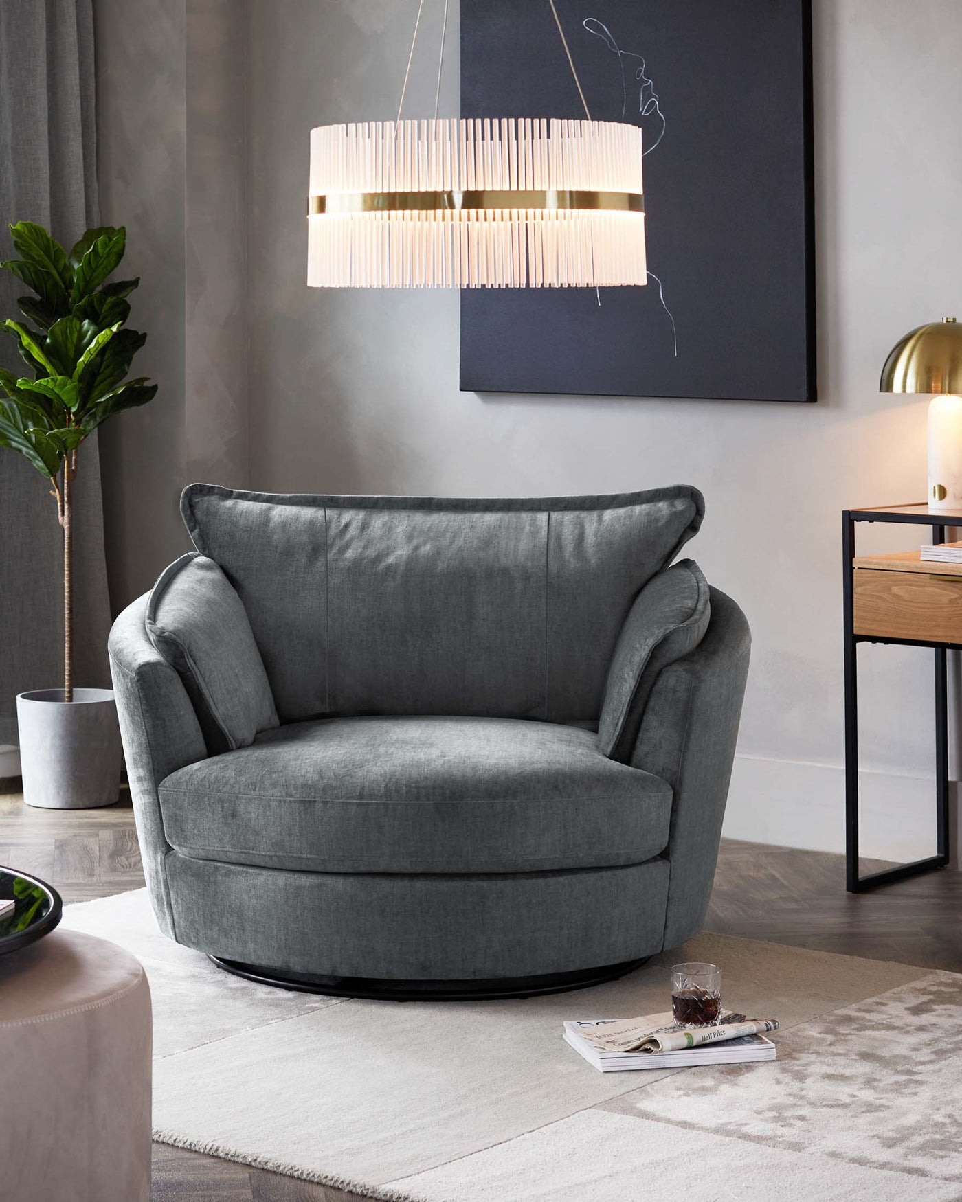 Plush, oversized round armchair in elegant grey fabric with a soft, deep seat and cushioned backrest, set on a low-profile black circular base. The chair is complemented by a textured off-white area rug beneath it and a sleek side table that partially visible to the right, displaying modern decor.