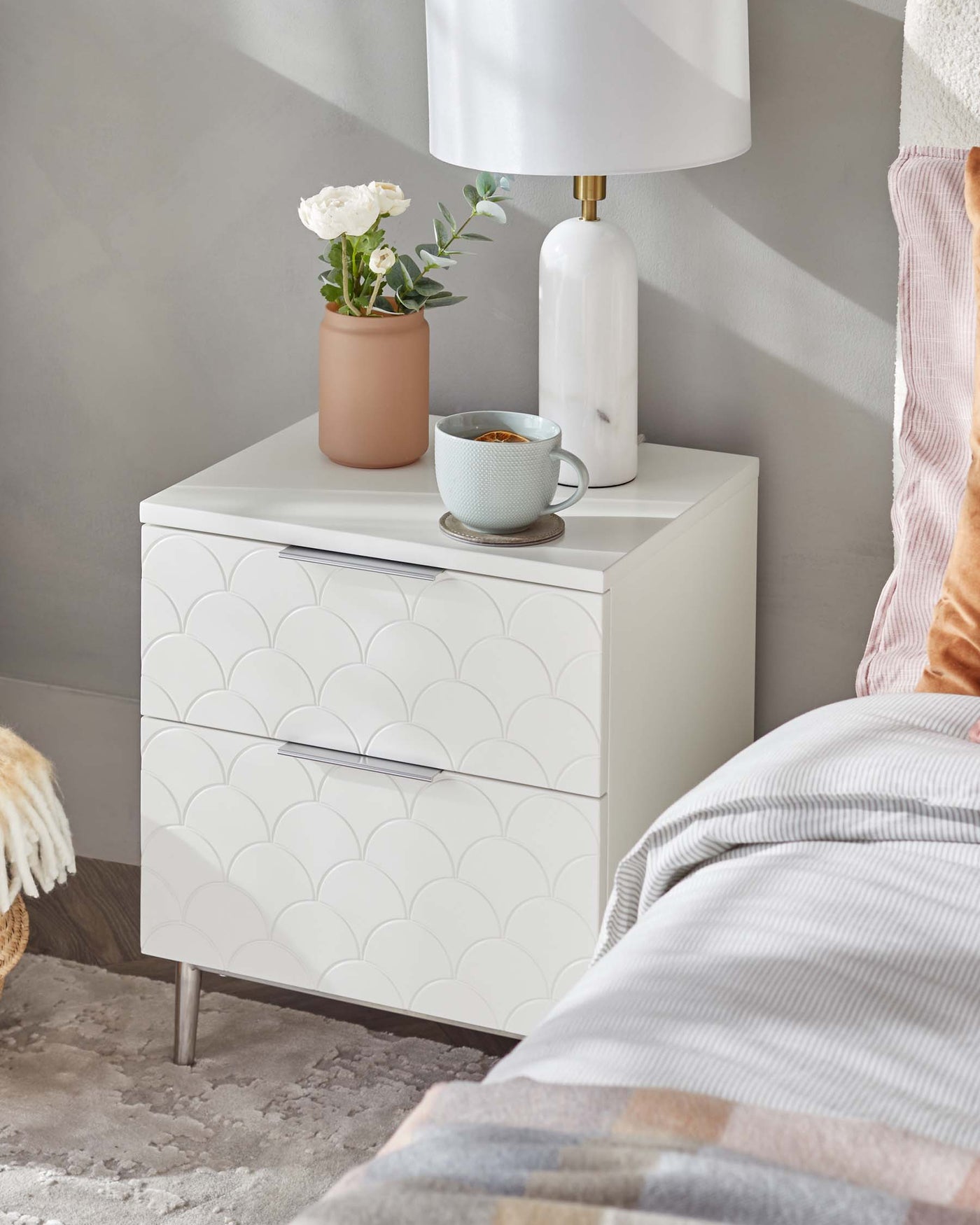 A contemporary white bedside table with a scallop patterned facade featuring two drawers with sleek silver handles, set against a grey wall and positioned next to a bed with pastel bedding.