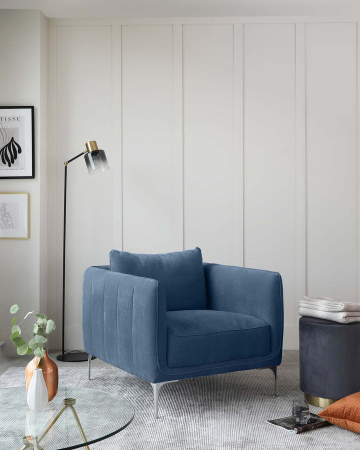 A contemporary blue velvet loveseat with vertical channel tufting and sleek metallic legs; a round glass-top side table with a gold-finished base; and a round, dark blue upholstered ottoman with a hidden storage feature and a brass base.