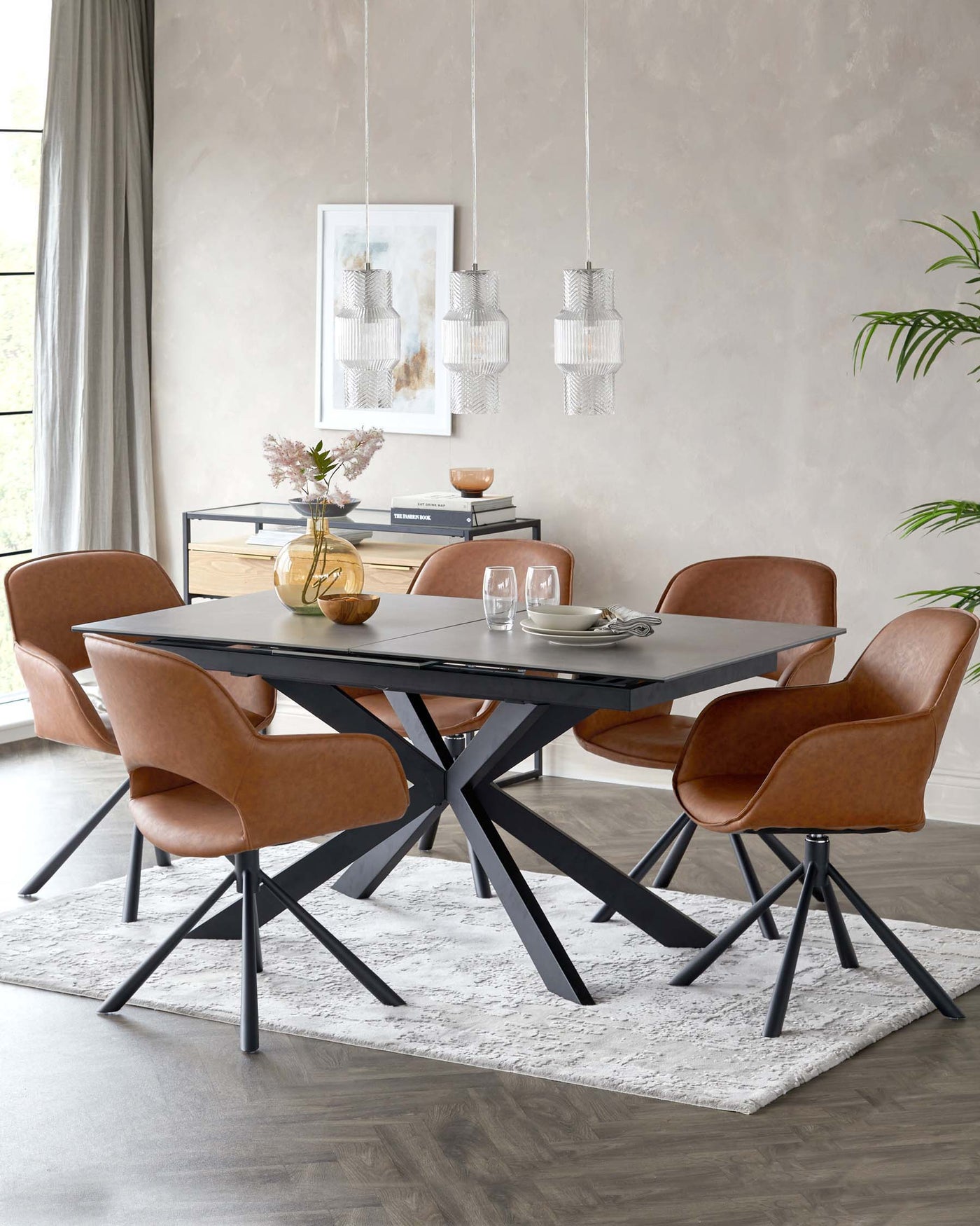Modern dining room set featuring a rectangular black table with a unique X-shaped base. Accompanied by six caramel brown upholstered chairs with black metal legs, situated on a textured white area rug. A minimalist sideboard with a combination of black metal and natural wood tones is positioned against the wall.