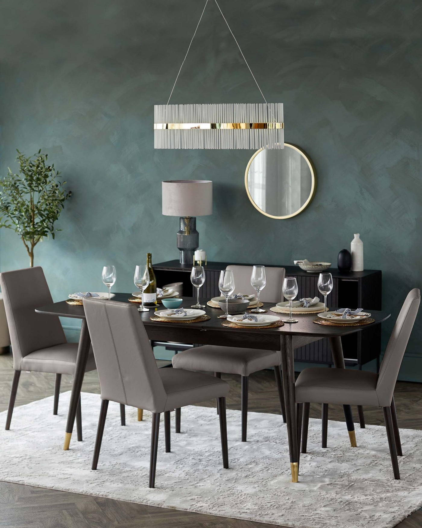 Elegant dining room setup featuring a modern rectangular wood dining table with gold-accented feet, surrounded by six matching taupe upholstered chairs with high backs and slim legs. A contemporary hanging pendant light with a linear design and metallic accents illuminates the space. A side cabinet and decorative round mirror with a thin gold frame complement the setting.