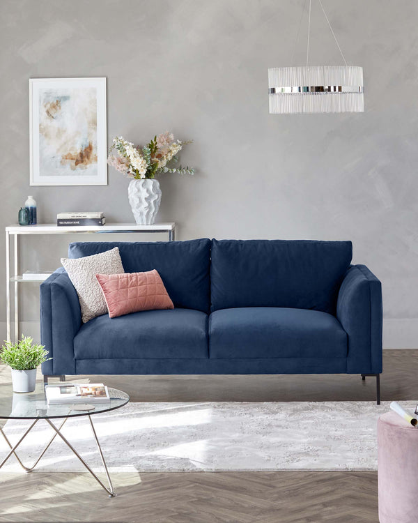 Navy blue upholstered three-seater sofa with plush cushions, white-painted wooden console table, round glass coffee table with a metal frame, light grey area rug, and a cylindrical pink fabric ottoman.