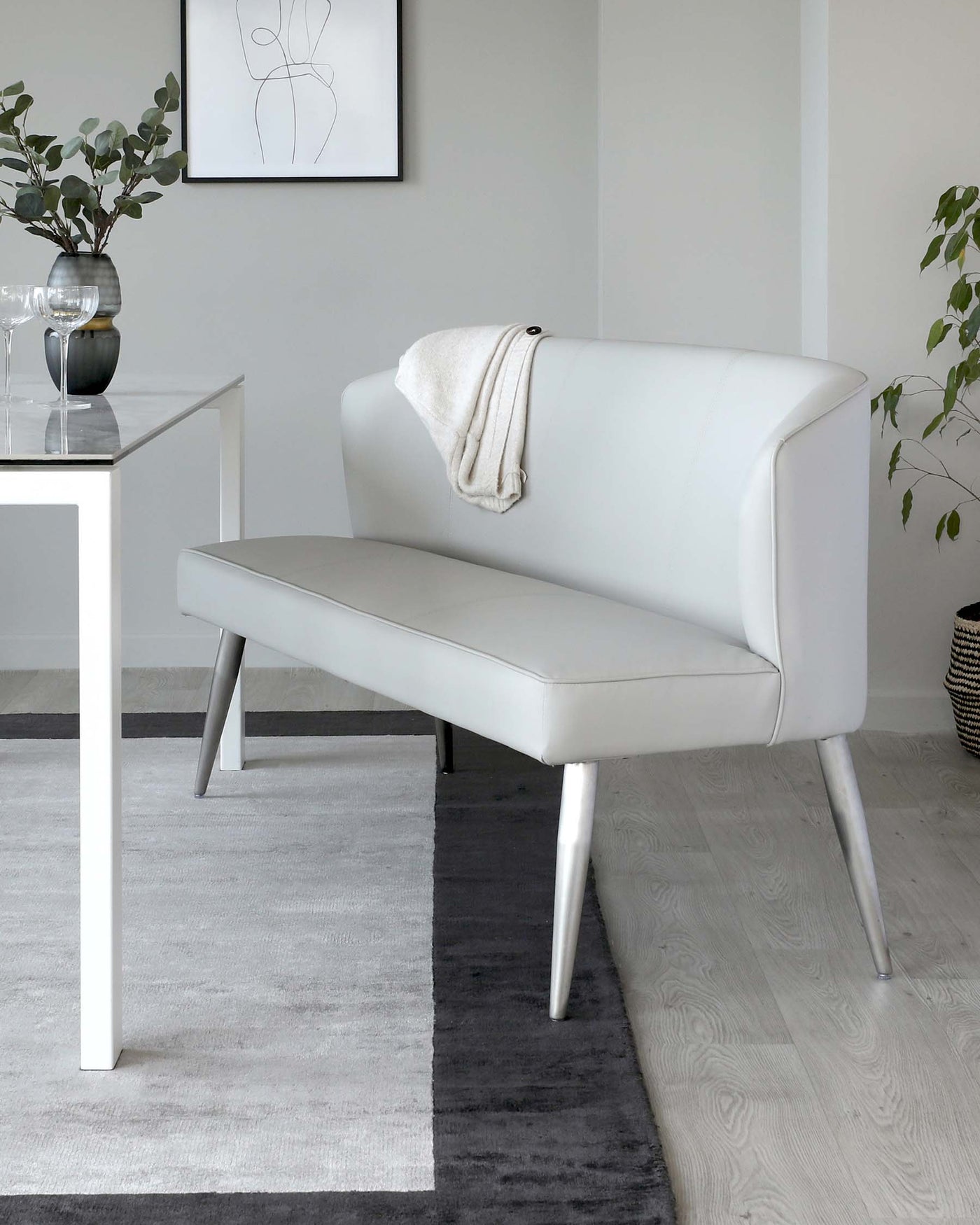 Elegant minimalist-style interior featuring a sleek white leather upholstered bench with a high backrest and angled metal legs, paired with a simple white square side table showcasing a glass vase and small plant. The setting is enhanced by a monochromatic area rug and subtle wall art, creating a modern and sophisticated space.
