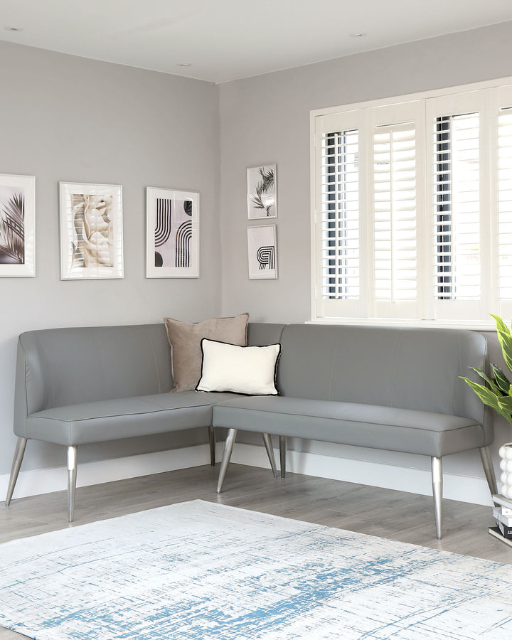 Modern corner sofa in a neutral grey with clean lines and metal legs, paired with a mix of solid and patterned cushions. A contemporary blue and white area rug lies beneath.