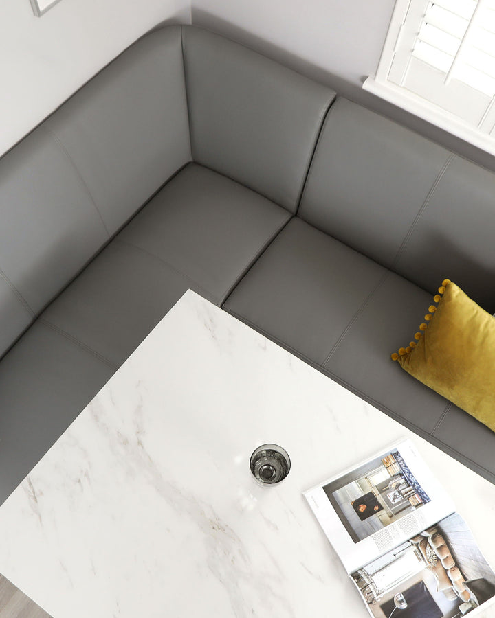 An L-shaped, grey upholstered bench seat with a built-in look, complemented by a squat, white marble-topped square coffee table with a central black vase. A mustard yellow throw pillow with pom-pom edges adds a pop of colour to the monochromatic setting. A lifestyle magazine is open on the table, suggesting a relaxed living space.