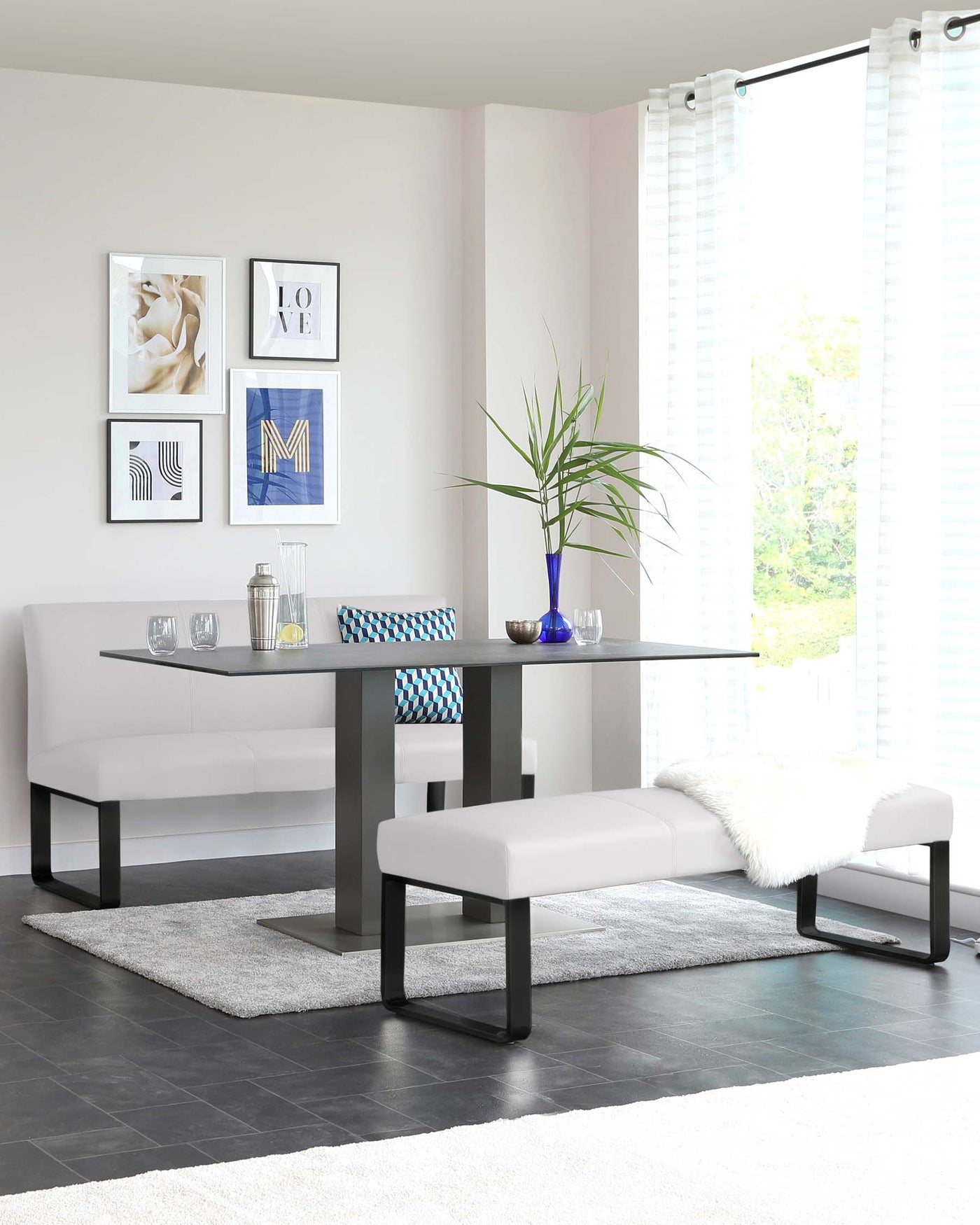 Modern minimalist dining furniture set featuring a sleek black dining table with a rectangular top and metal base paired with a matching bench and a padded backless bench. A white fluffy throw is draped over one end of the backless bench, complementing the light neutral colour scheme of the room.