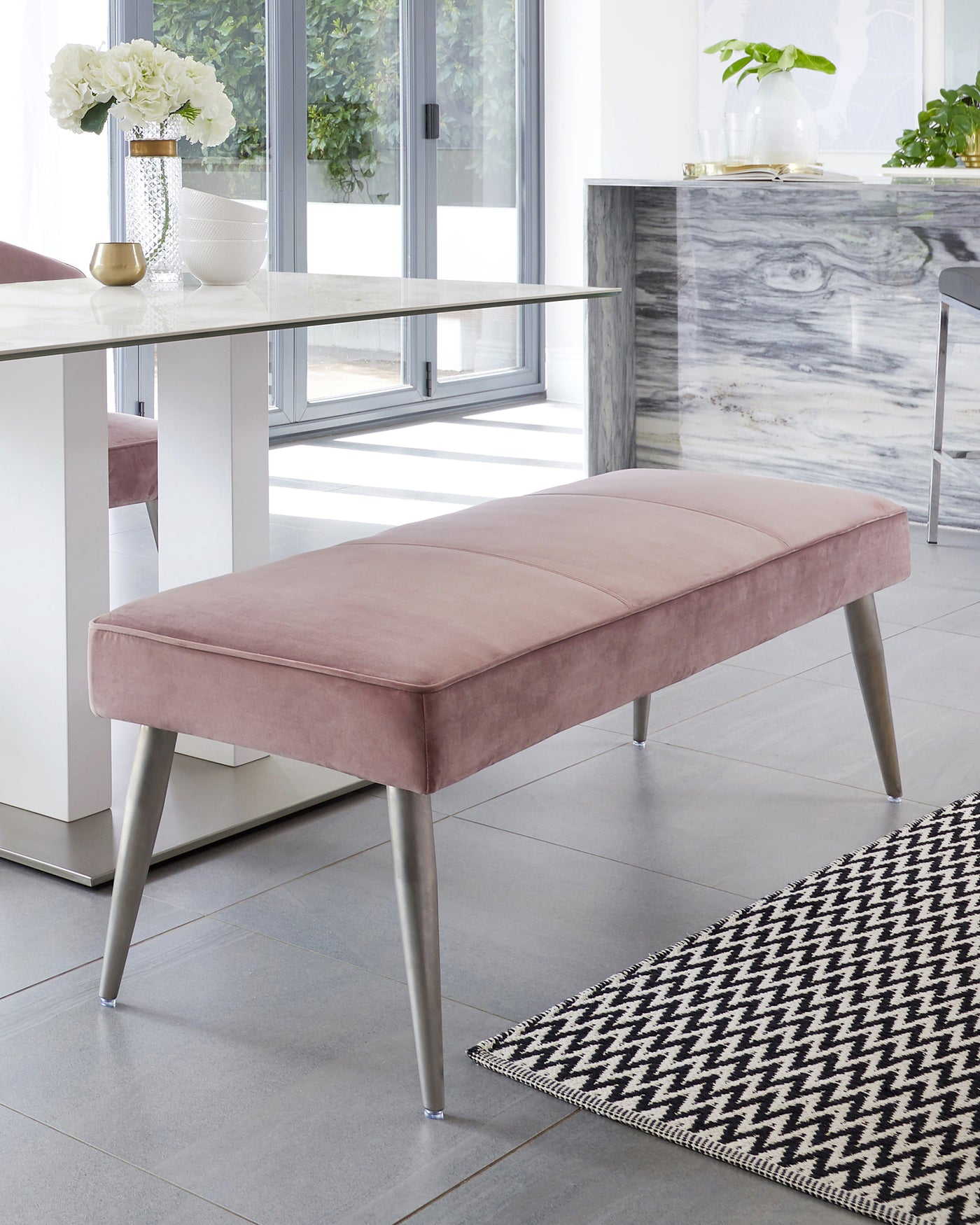 Modern sleek bench with a plush pink velvet cushion and tapered metallic legs positioned in a bright, contemporary room.