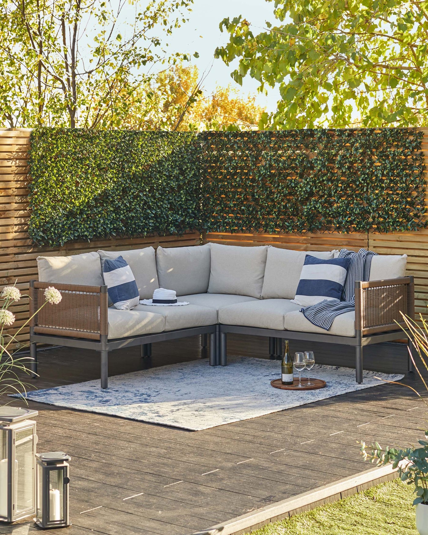 Outdoor corner sectional sofa with beige cushions, wicker accents, and a deep brown frame. The sectional is accessorized with navy and white striped throw pillows. A light-coloured outdoor rug lies beneath the sofa, and a small round wooden tray table with a bottle and glasses sits on the rug. Two metal lanterns rest to the side on the wooden deck amidst a garden setting with lush greenery.