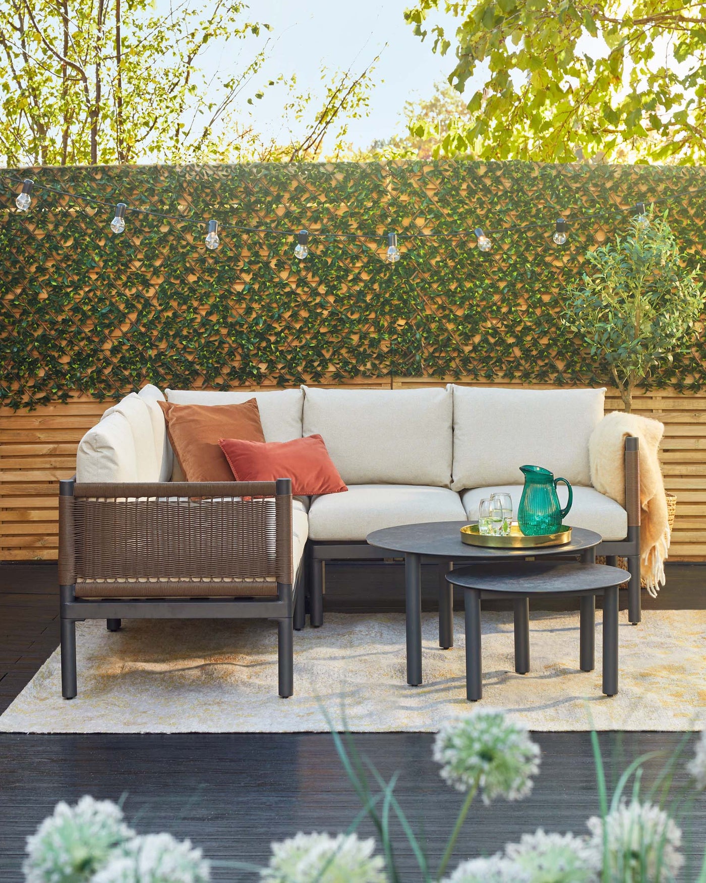 Modern outdoor furniture set including a beige cushioned corner sectional sofa with accent pillows, a matching armchair with wicker back, and two round nested coffee tables in black finish on a patterned area rug.