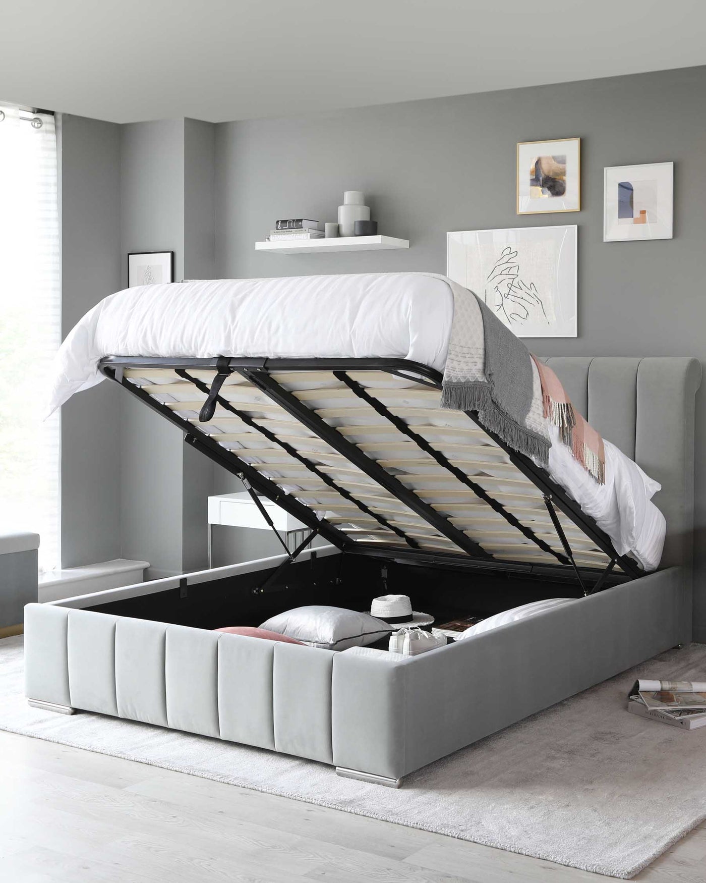 Gray upholstered storage bed with a lift-up frame revealing a spacious under-mattress storage compartment. The bed features a padded, channel-tufted headboard and is dressed with white and grey bedding. It rests on a light grey area rug, and the bed's modern design is complemented by minimalist wall art and a sleek white floating shelf above.