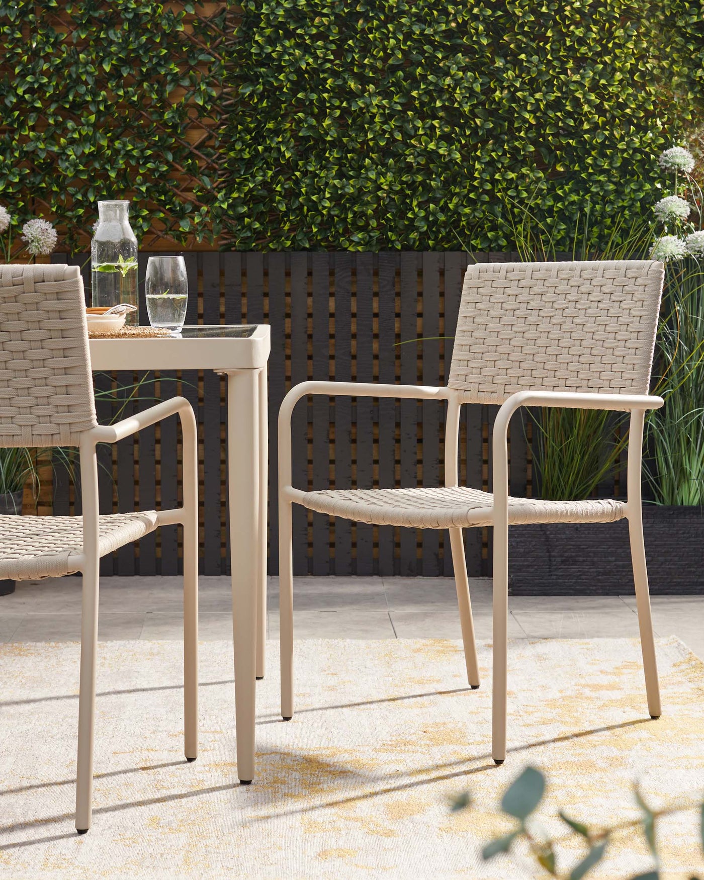 Outdoor furniture featuring two modern beige armchairs with woven backrests and seats, and a matching slim beige square table with a clear vase and glass set upon it, situated on a textured cream and yellow area rug. The scene is complemented by a backdrop of a green leafy hedge and a dark wooden slat fence, evoking an inviting and contemporary patio ambiance.