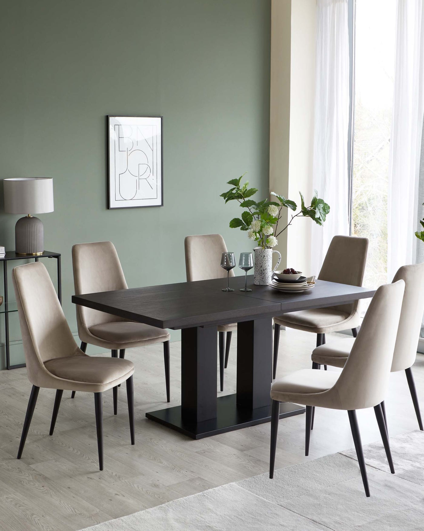 Elegant dining room featuring a modern rectangular dark wood table with a sleek, black base, surrounded by six taupe upholstered chairs with slim black legs. To the left, a matching dark wood sideboard with glass doors hosts decorative items and a grey lamp with a round shade.