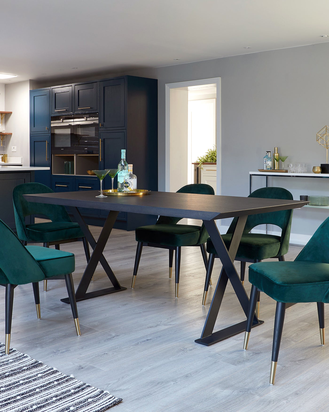 Modern dining room furniture featuring a dark wood table with a rectangular top and angular metal legs, accompanied by six plush, green velvet chairs with black tapered legs and gold feet caps. A minimalist black sideboard with kitchen cabinets in the background completes the scene.
