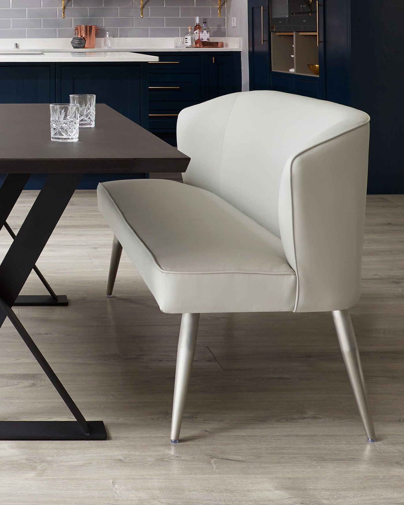 Modern minimalist dining chair with beige leather upholstery and sleek, slender metal legs. The chair features a curved backrest for comfortable seating, complementing a dark wooden dining table in the background.