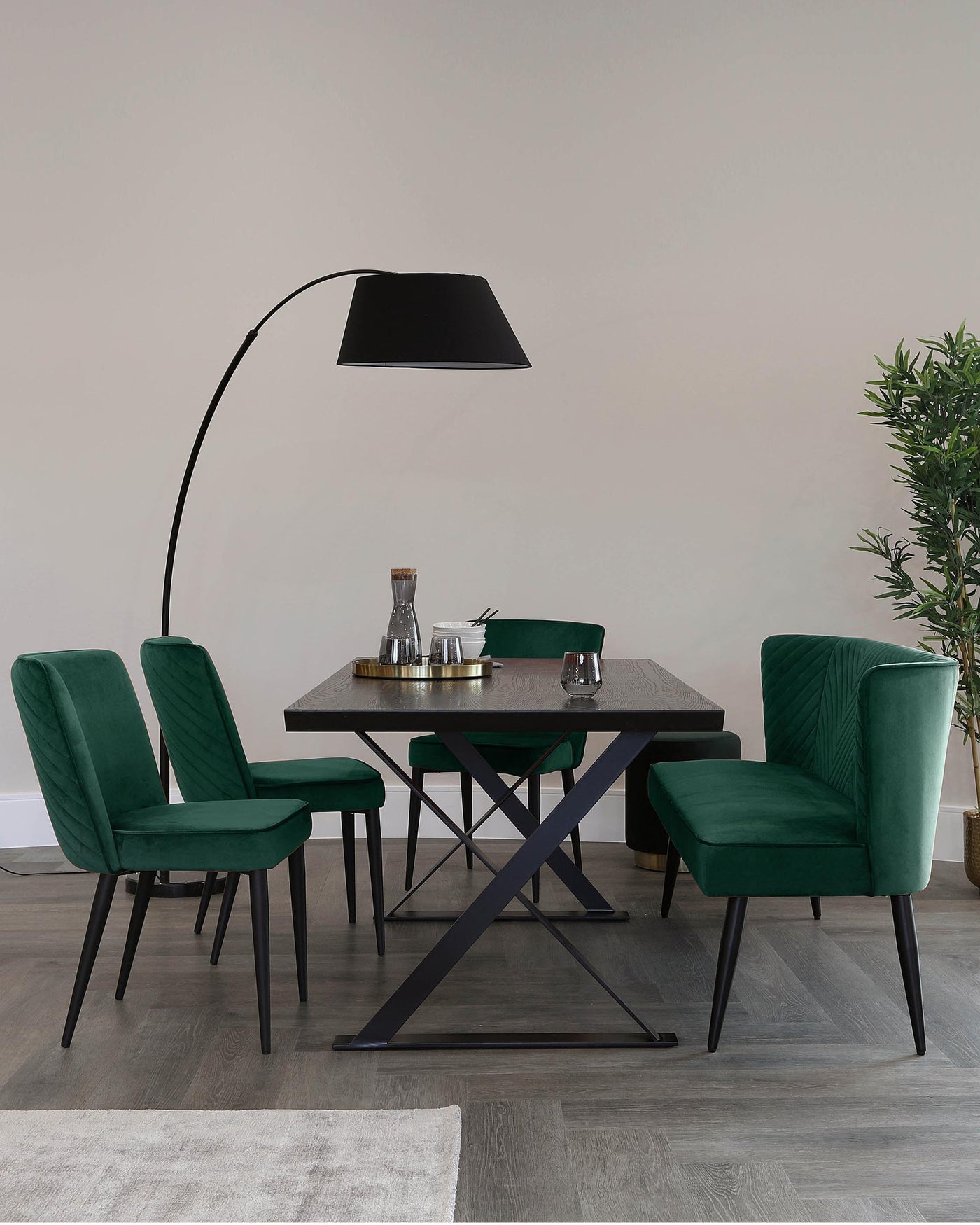Modern dining room furniture set featuring a dark wood table with a unique X-shaped black metal base and four plush green velvet chairs with vertical stitch detailing and black metal legs. An arched floor lamp with a large black shade extends over the table, creating a contemporary atmosphere.