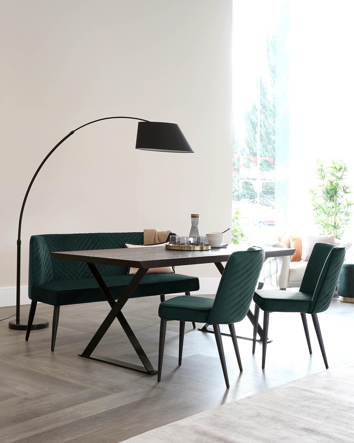 Modern dining room with a dark wooden table with an X-shaped metal base, surrounded by three teal upholstered dining chairs with black metal legs. An arched floor lamp with a large black shade looms over the setup.