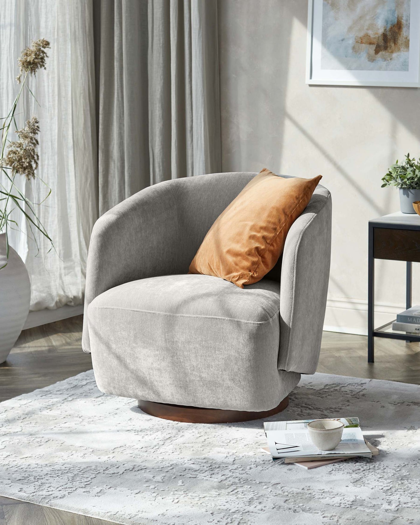Modern light grey fabric barrel chair with plush cushioning and a golden-brown velvet accent pillow, set on a natural wood circular base. Positioned on a textured white area rug beside a small dark wood side table with books and a coffee cup, in a room with light grey curtains and abstract wall art.