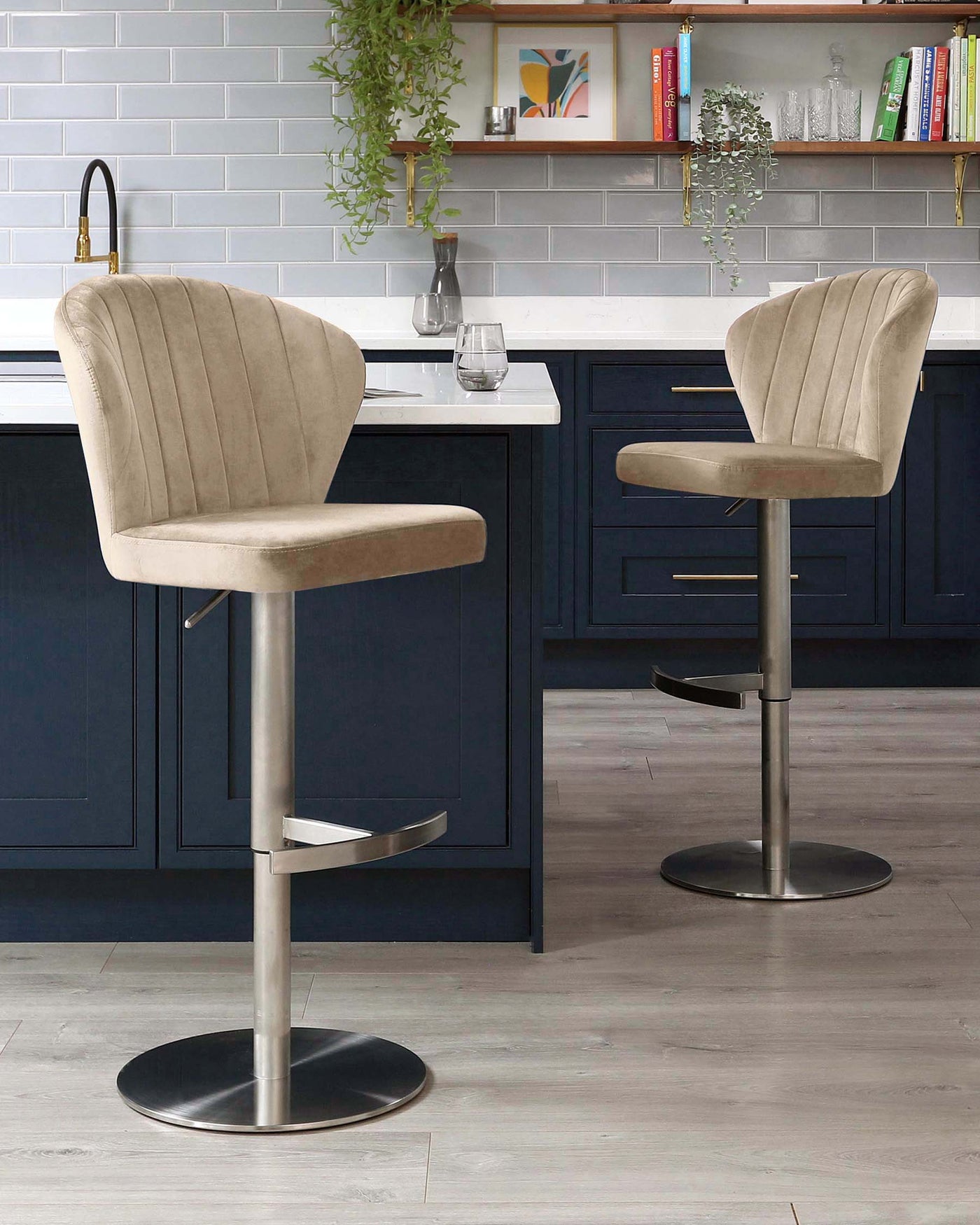 Two contemporary upholstered bar stools with vertical channel tufting, soft beige fabric, and a sleek pedestal base in a chrome finish, featuring a footrest and adjustable height function. The stools are positioned at a kitchen island with a blue cabinet and white countertop, complemented by a grey subway tile backsplash.