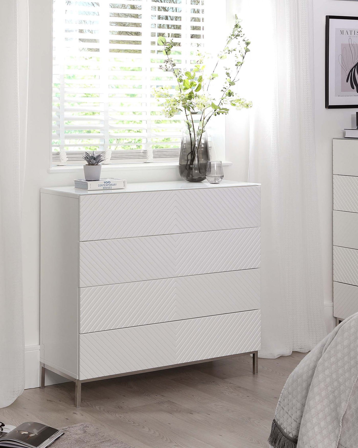 Modern white chest of drawers with a textured herringbone pattern on the fronts, complemented by sleek metallic handles and legs, staged in a bright room with natural light filtering through window shutters.