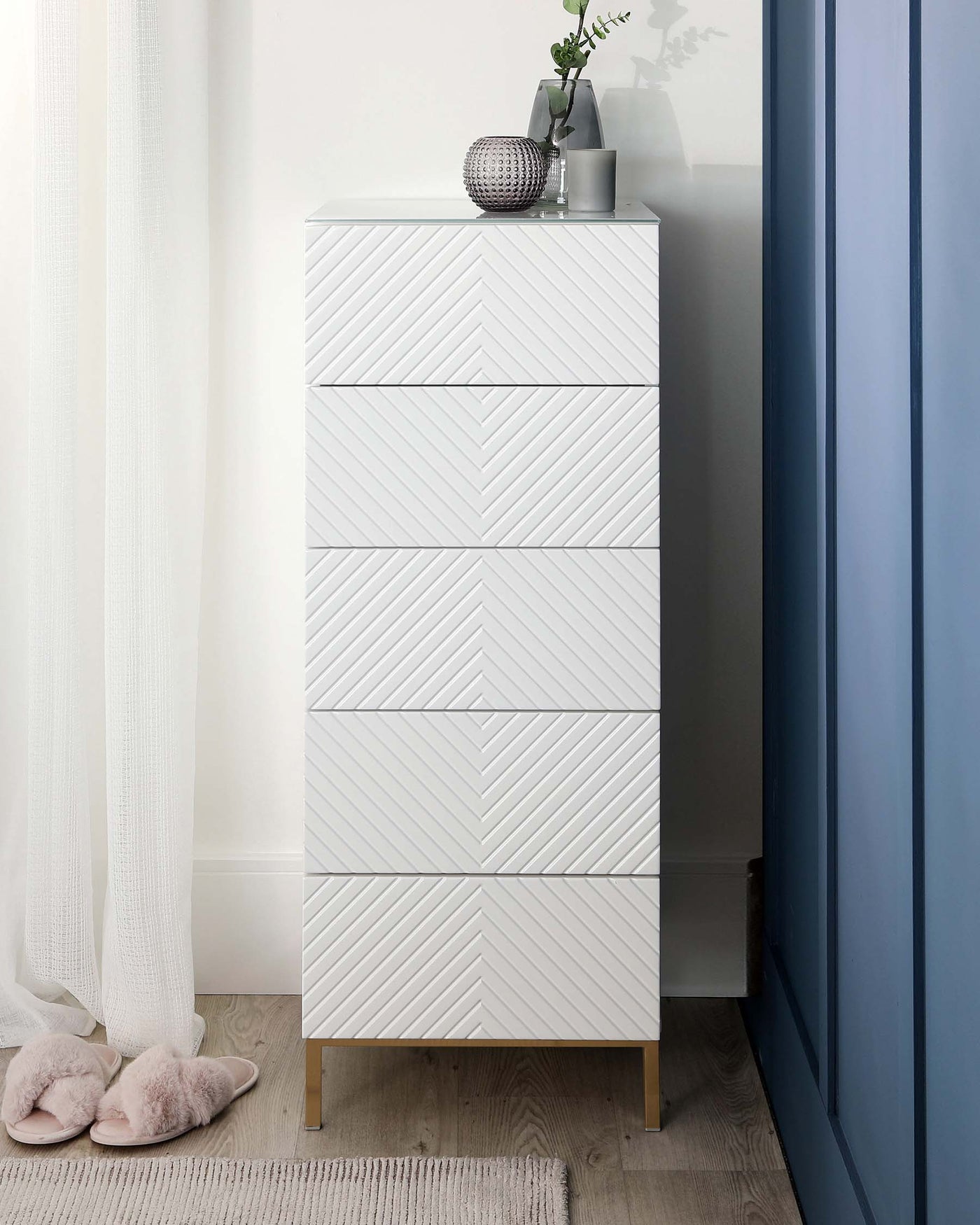 A modern white tallboy chest of drawers with a textured chevron pattern on the drawer fronts and light wooden legs. The dresser is accessorized with a decorative vase, green plant, and a textured sphere atop its smooth surface. The furniture piece stands against a light-coloured wall beside a blue curtain and a white sheer, with a cosy pair of slippers on the floor and a woven mat.