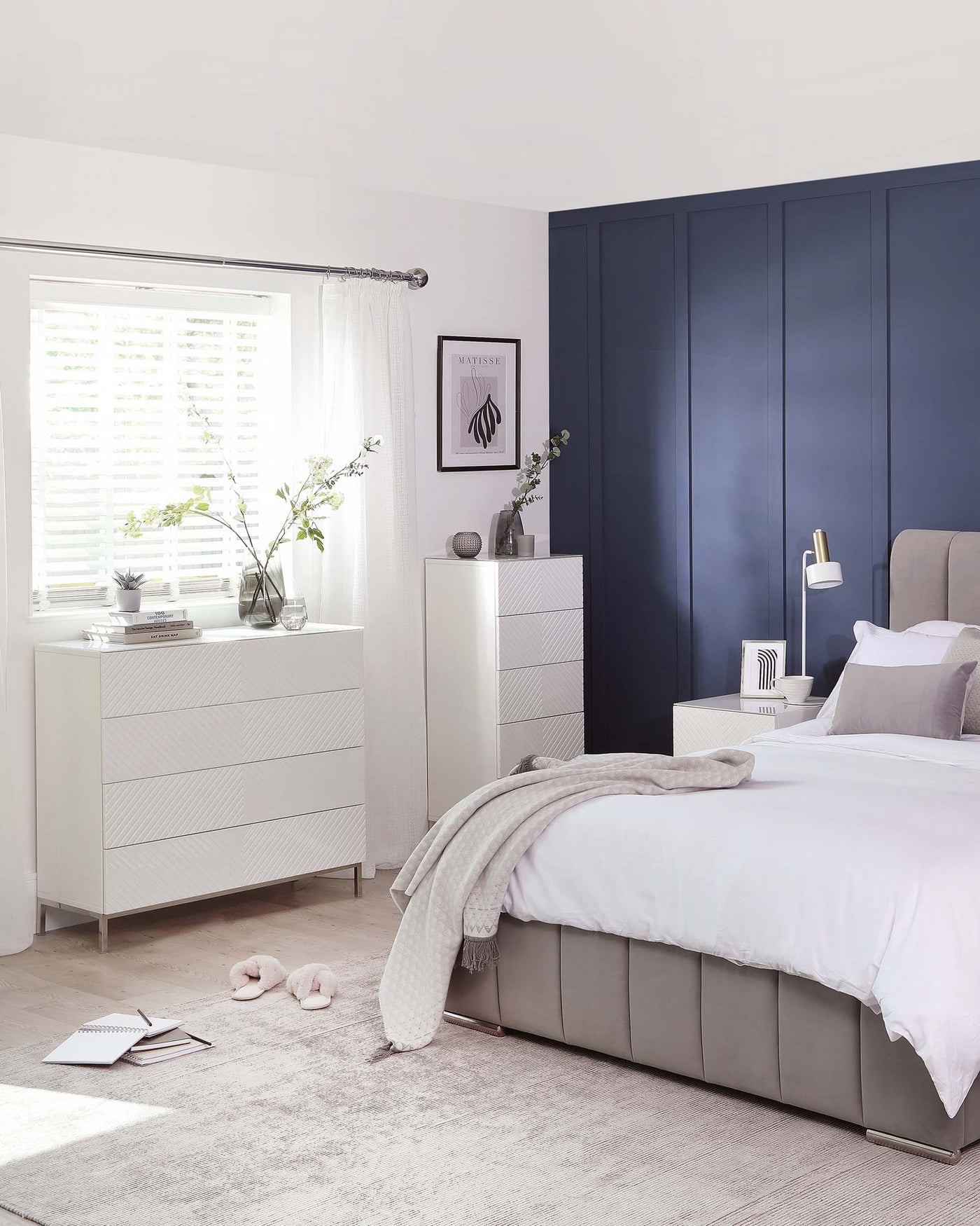 Modern bedroom furniture including a grey upholstered bed with a high headboard, a white textured dresser, and a matching bedside table. The dresser and bedside table have a geometric carved design and metal accents.