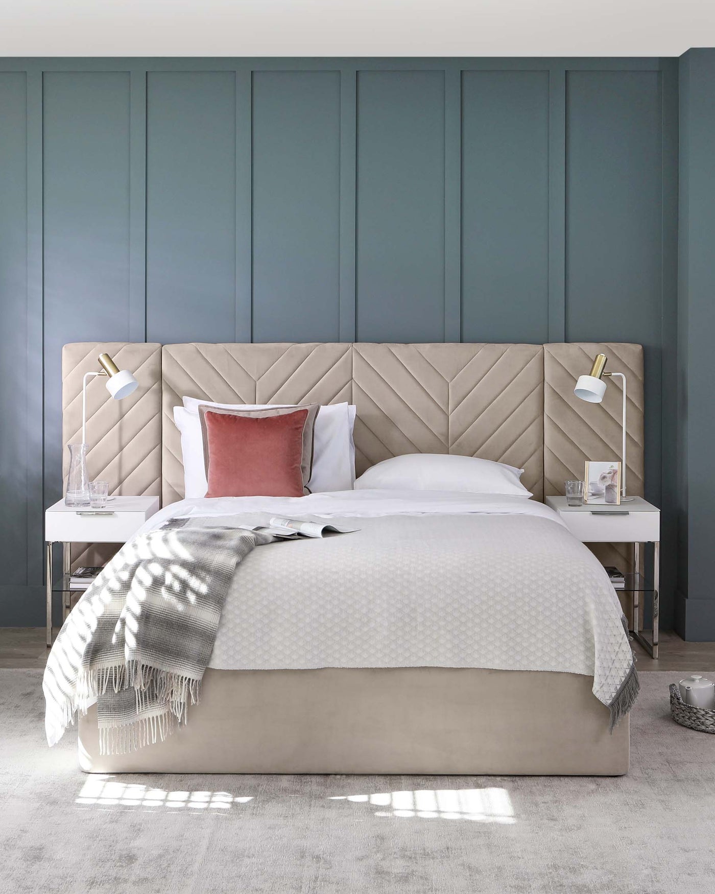 Elegant upholstered bed with an extended, tufted headboard in a herringbone pattern, flanked by two white, minimalist nightstands with gold-accented reading lamps. Bed is adorned with crisp white linens, a textured white comforter, and accented by a single dusty rose pillow. A grey and white patterned throw blanket casually drapes over the foot of the bed. The bed and nightstands are set against a room with dark teal panelled walls and a soft grey carpet.