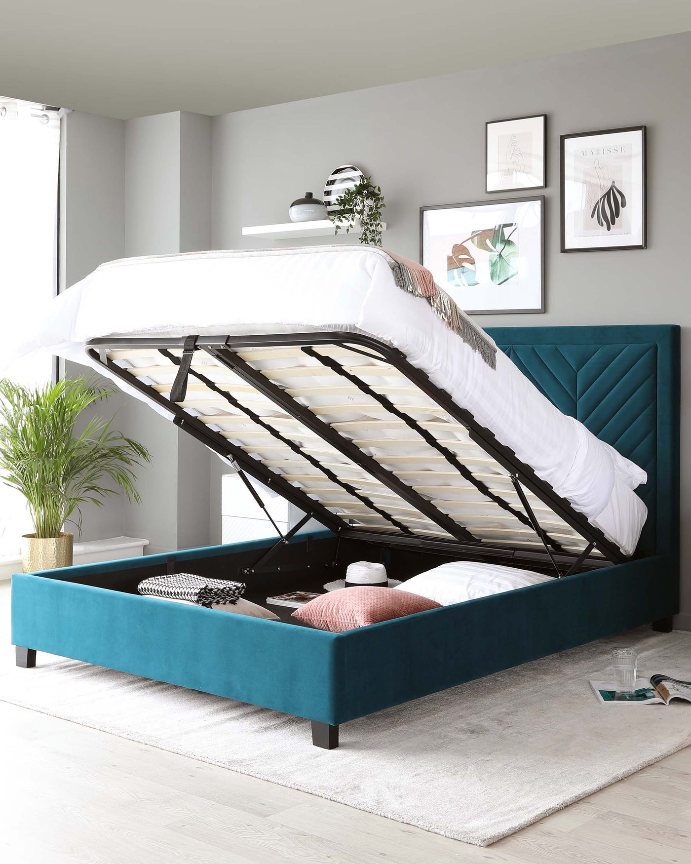 A contemporary teal upholstered storage bed with a lifted mattress base revealing internal storage, set against a neutral grey wall in a well-lit room. The headboard features a tufted vertical channel design, and the bed stands on dark wooden legs. The bedding includes white linens partially draped aside, and nearby accent pillows rest inside the open storage space. A potted plant adds a touch of greenery to the space.