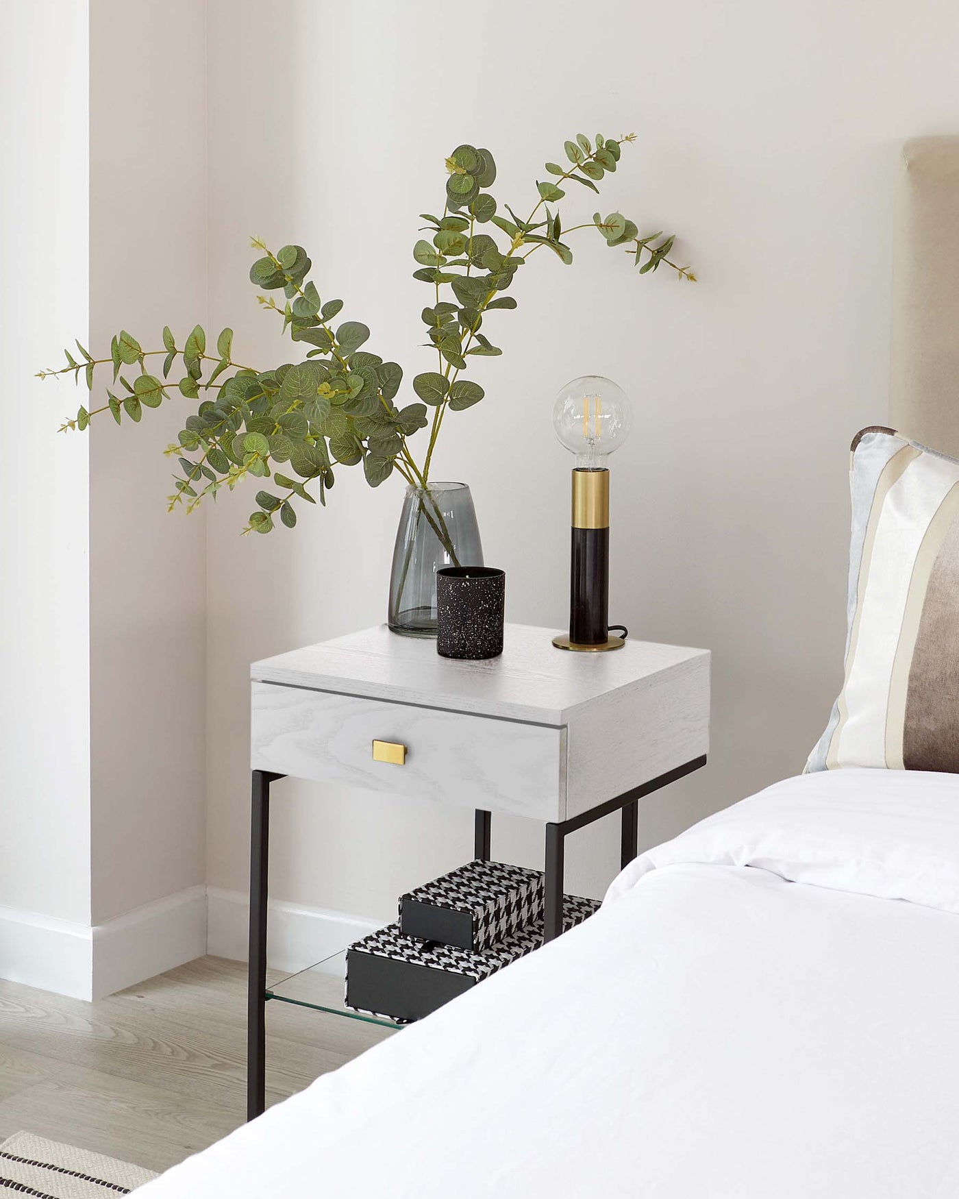 Modern bedside table with a faux marble finish on top, featuring a single drawer with a brass-coloured handle, supported by a minimalist black metal frame and including a lower glass shelf.