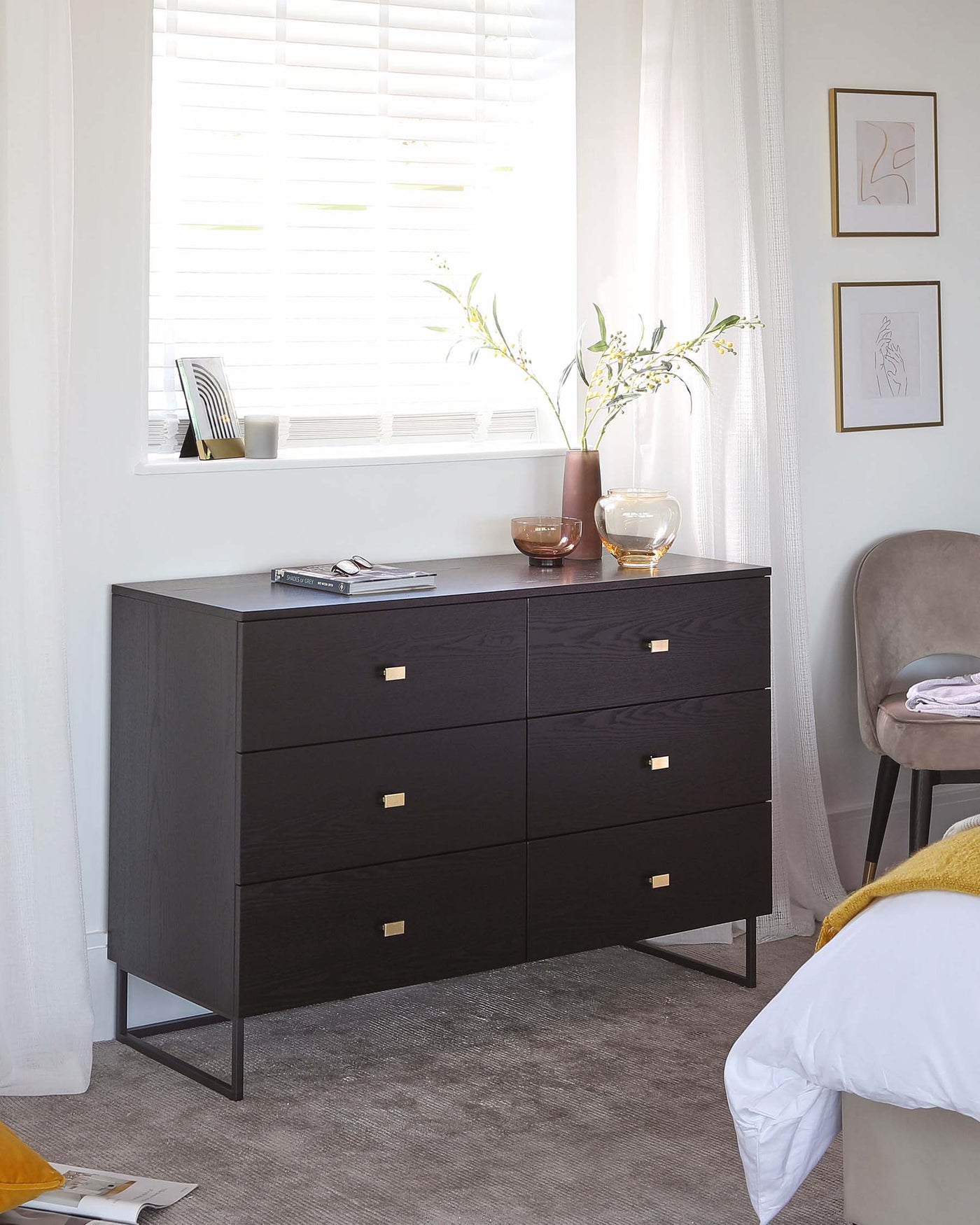 Modern black wooden dresser with six drawers featuring brass rectangular handles, standing on sleek metal legs. There are decorative items on top, including vases and books, complemented by a framed artwork on the wall above. A grey upholstered chair is partially visible to the right.