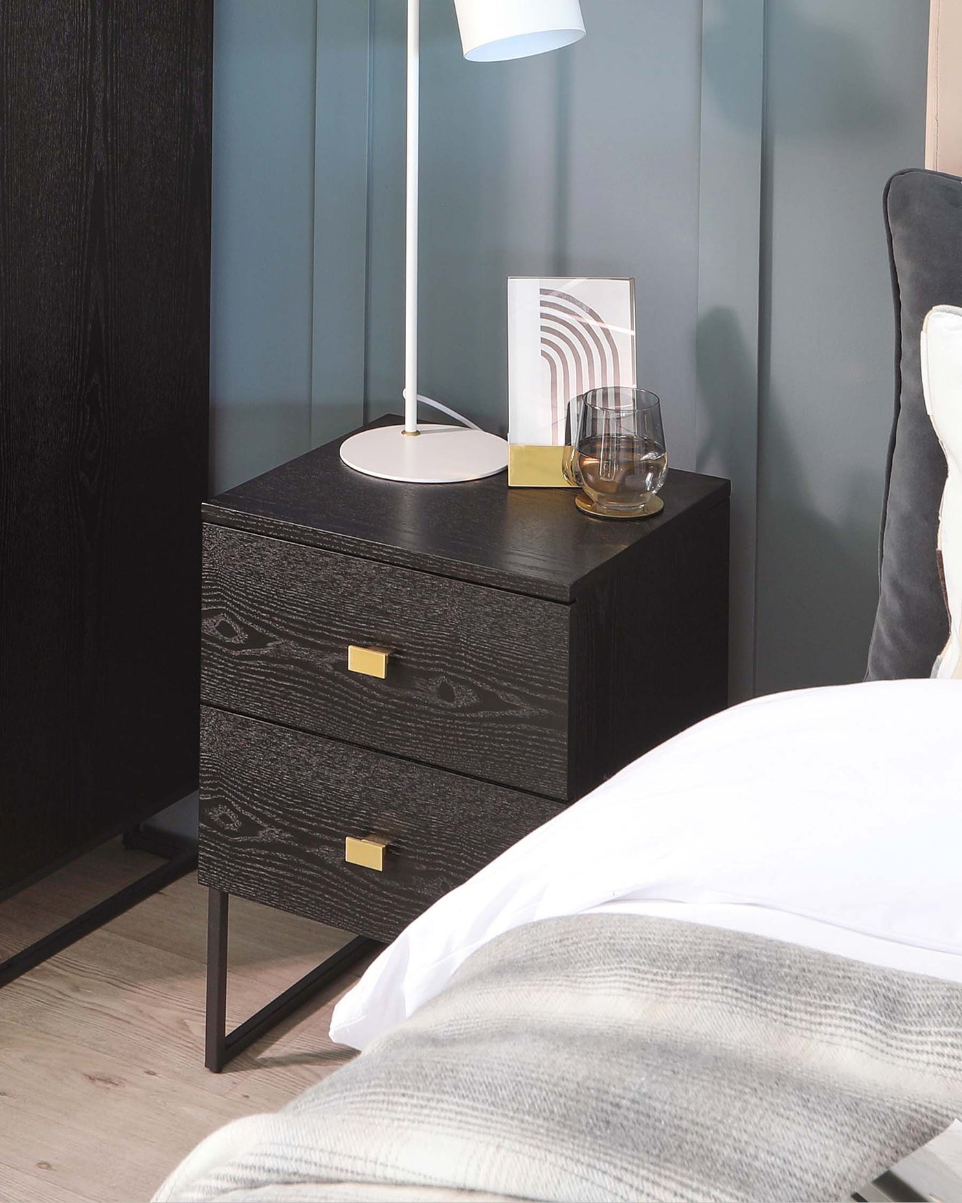 Modern black wood-grain bedside table with two drawers featuring sleek gold handles, positioned on metal legs against a teal wall, accompanied by a tall white floor lamp, decorative framed artwork, and a clear glass atop.
