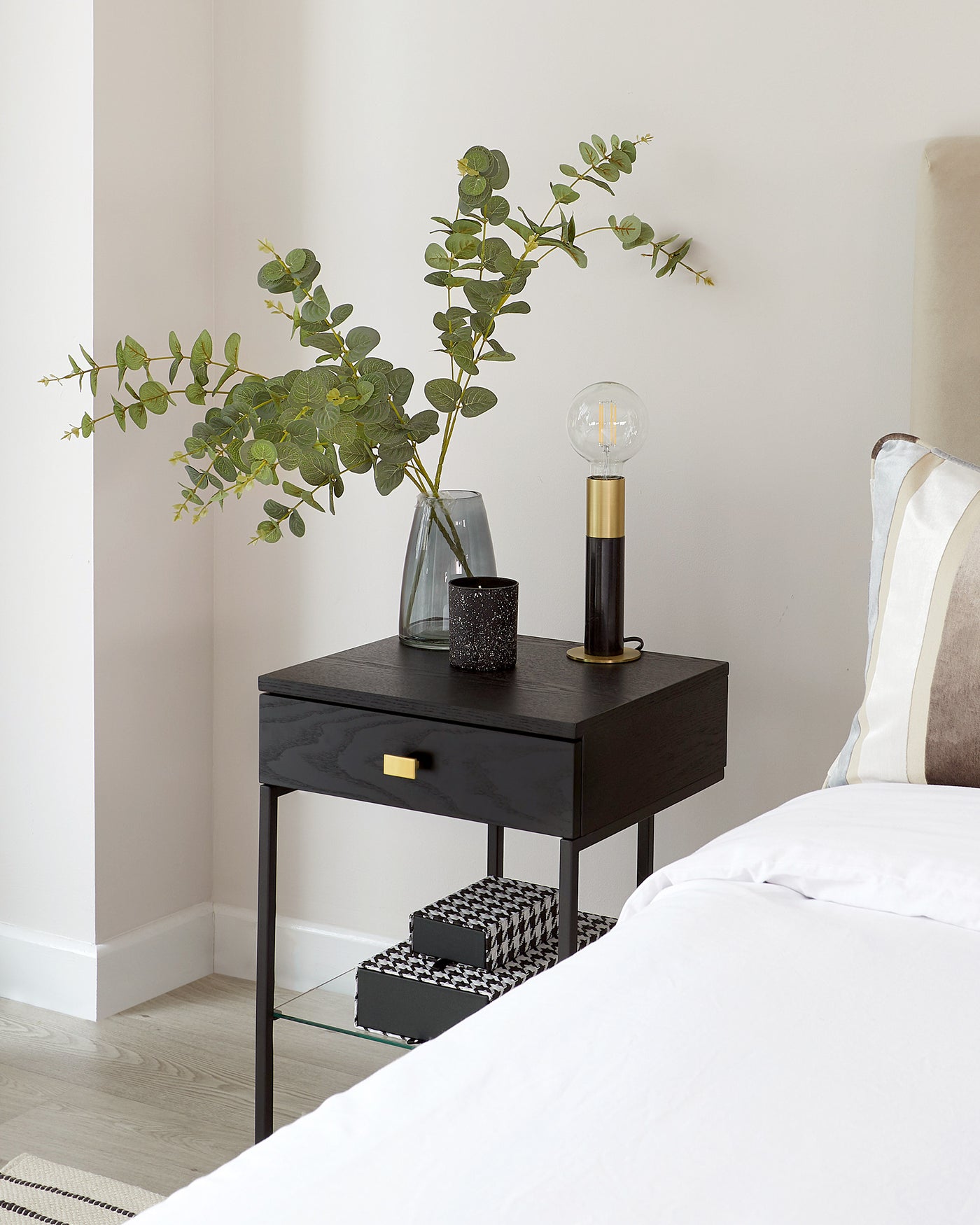 Elegant modern black bedside table with a sleek finish, featuring a single drawer with a brass pull handle, and a lower shelf with a glass insert. The table is styled with a contemporary glass vase with green branches, a decorative black textured candle holder, and a unique table lamp with a clear glass bulb and brass base.