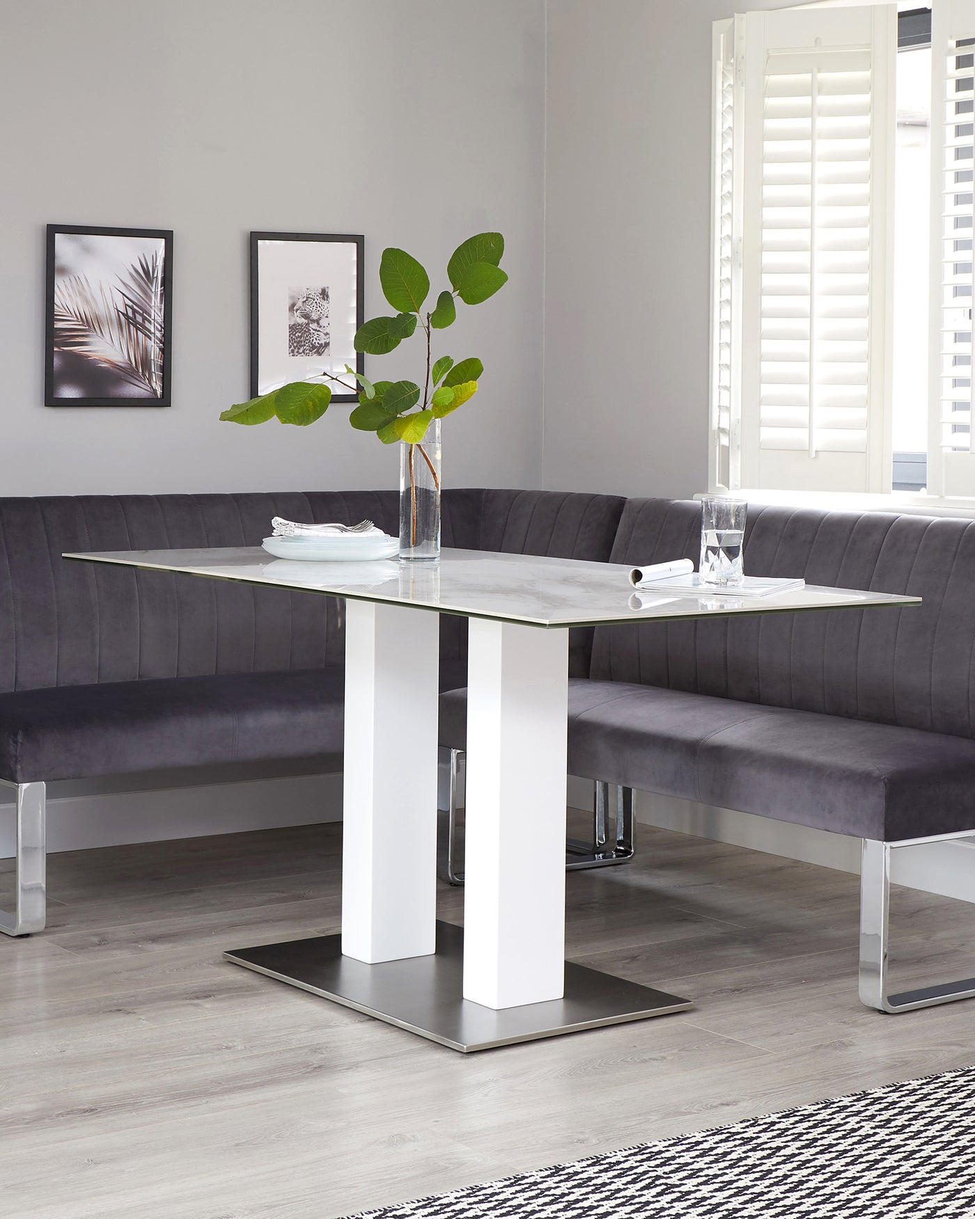 Modern dining room furniture featuring a white marble-topped table with rectangular surface, supported by two wide, white rectangular columns on a flat stainless-steel base. A matching corner bench upholstered in a tufted grey fabric provides comfortable seating along two walls.