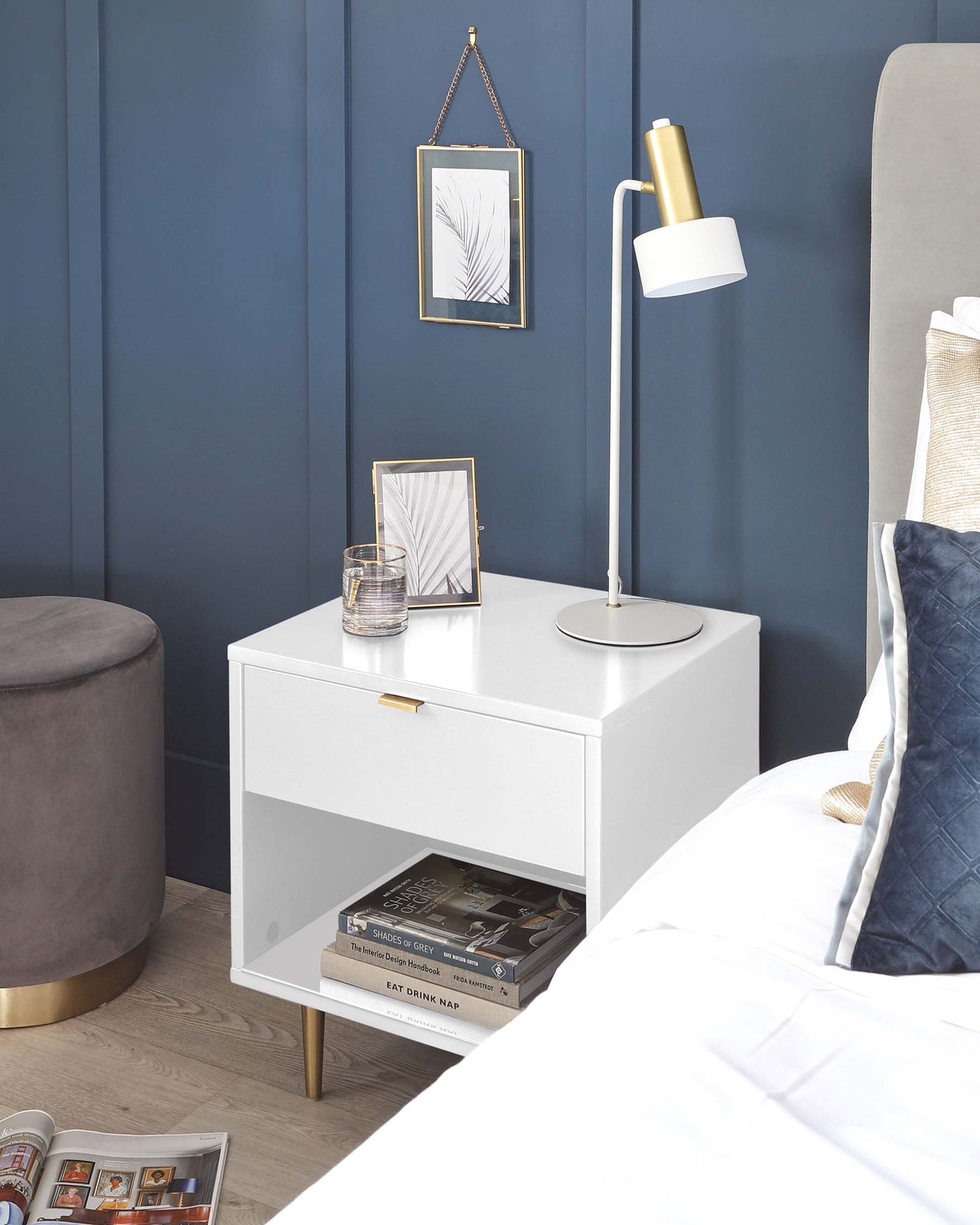 Modern white bedside table with a sleek design, featuring a single drawer with a gold-coloured handle, an open lower shelf with space for books, and a tabletop supporting various decorative items. A round upholstered ottoman with a velvet finish and gold base is partially visible to the left. The pieces exude contemporary elegance and functionality.