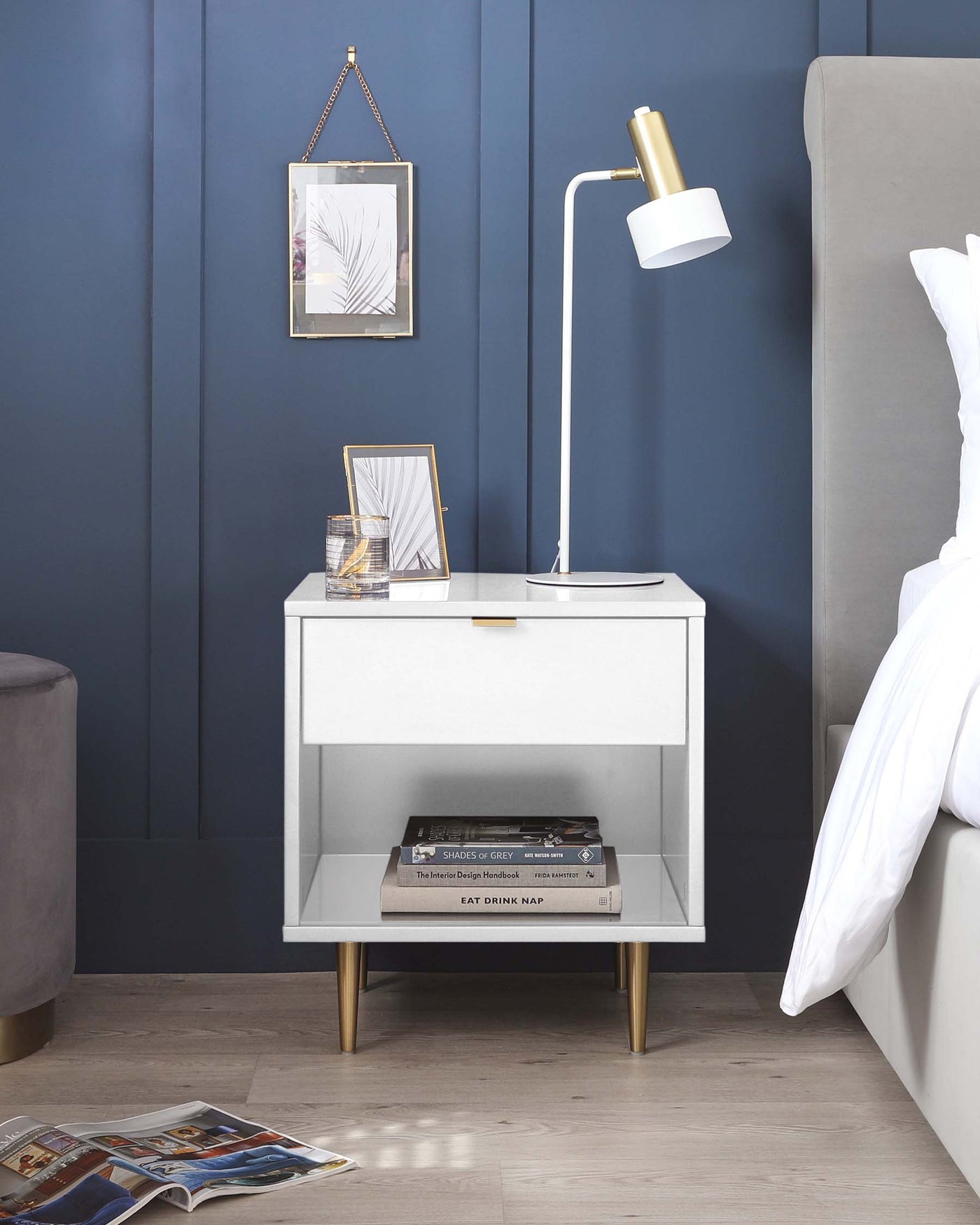 Modern white bedside table with a single drawer, gold handles, and gold-tipped legs, featuring an open lower shelf with stacked books. The table is accessorized with a white and gold desk lamp, framed pictures, and a decorative item, set against a blue wall with a hung framed artwork.