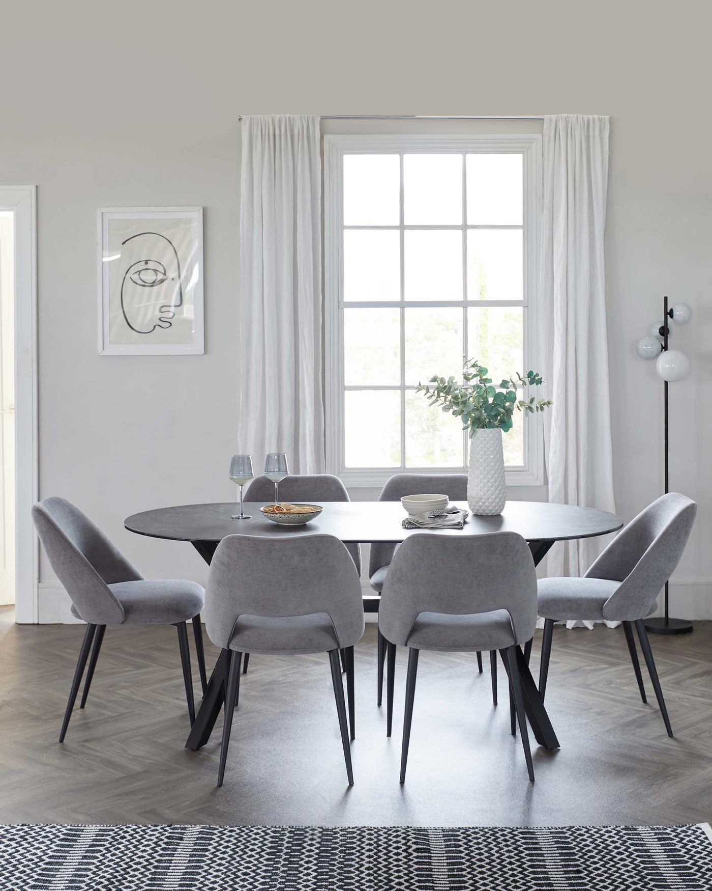 A modern dining area featuring a circular table with a dark stone top and black angular legs, surrounded by six plush, light grey upholstered chairs with tapered black legs. A stylish white vase with green foliage sits atop the table. The setting is complemented by a geometric black and white area rug beneath.