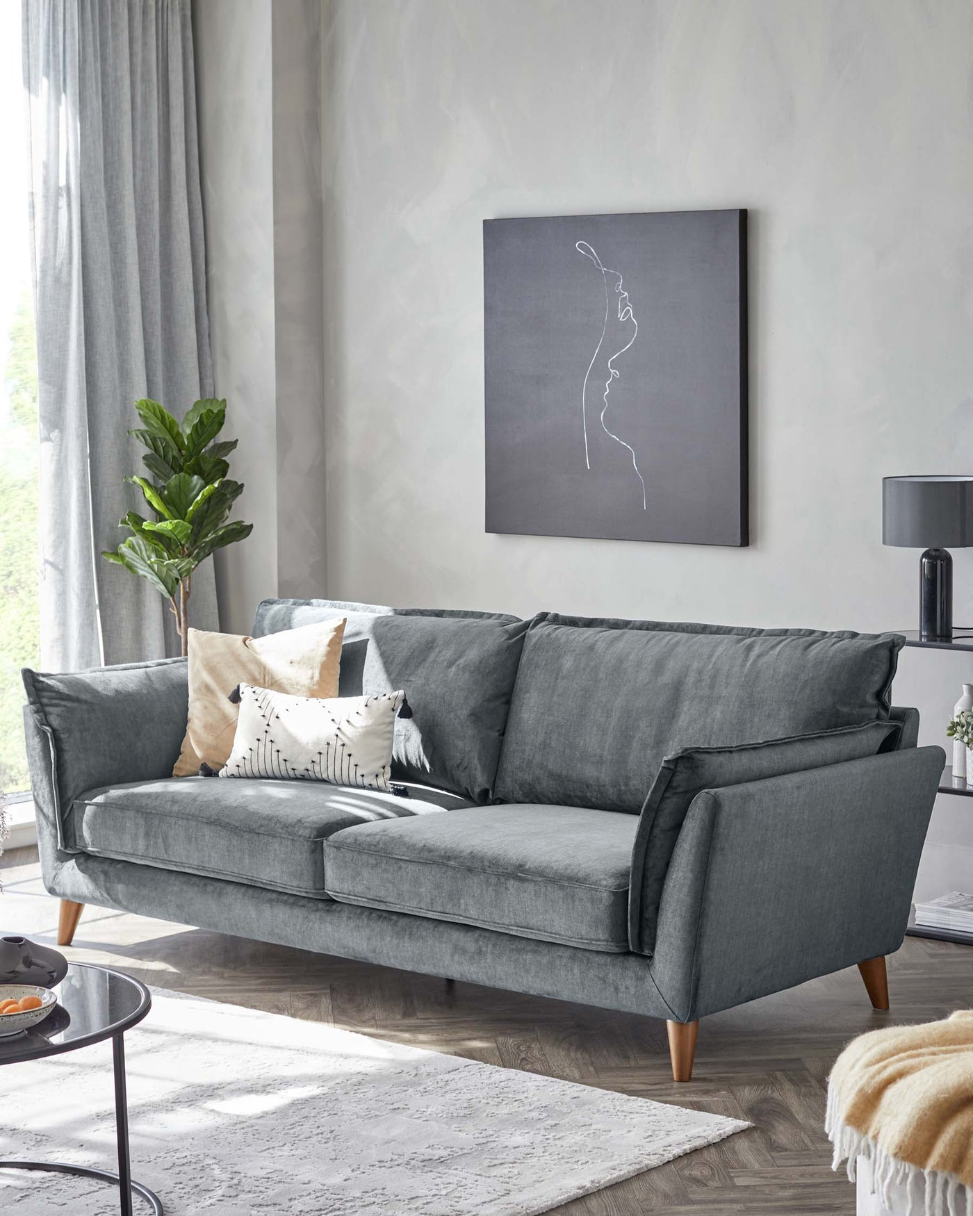 A contemporary three-seater sofa with plush grey fabric upholstery, featuring a tufted backrest, clean lines, and wooden legs. The sofa is accessorized with assorted throw pillows in neutral tones. In the forefront, there's a round, black side table with a minimalistic design.