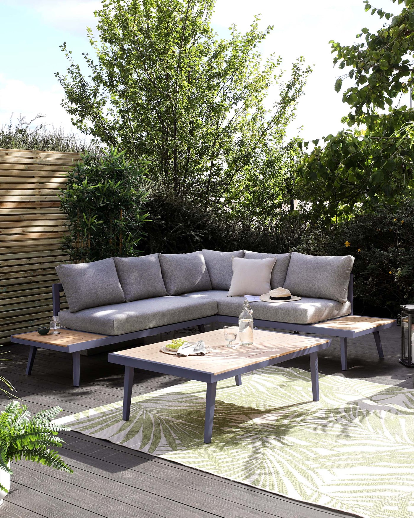 Modern outdoor sectional sofa set with grey cushions on a wooden frame, accompanied by two matching wooden coffee tables with grey legs, staged on an open patio surrounded by lush greenery.