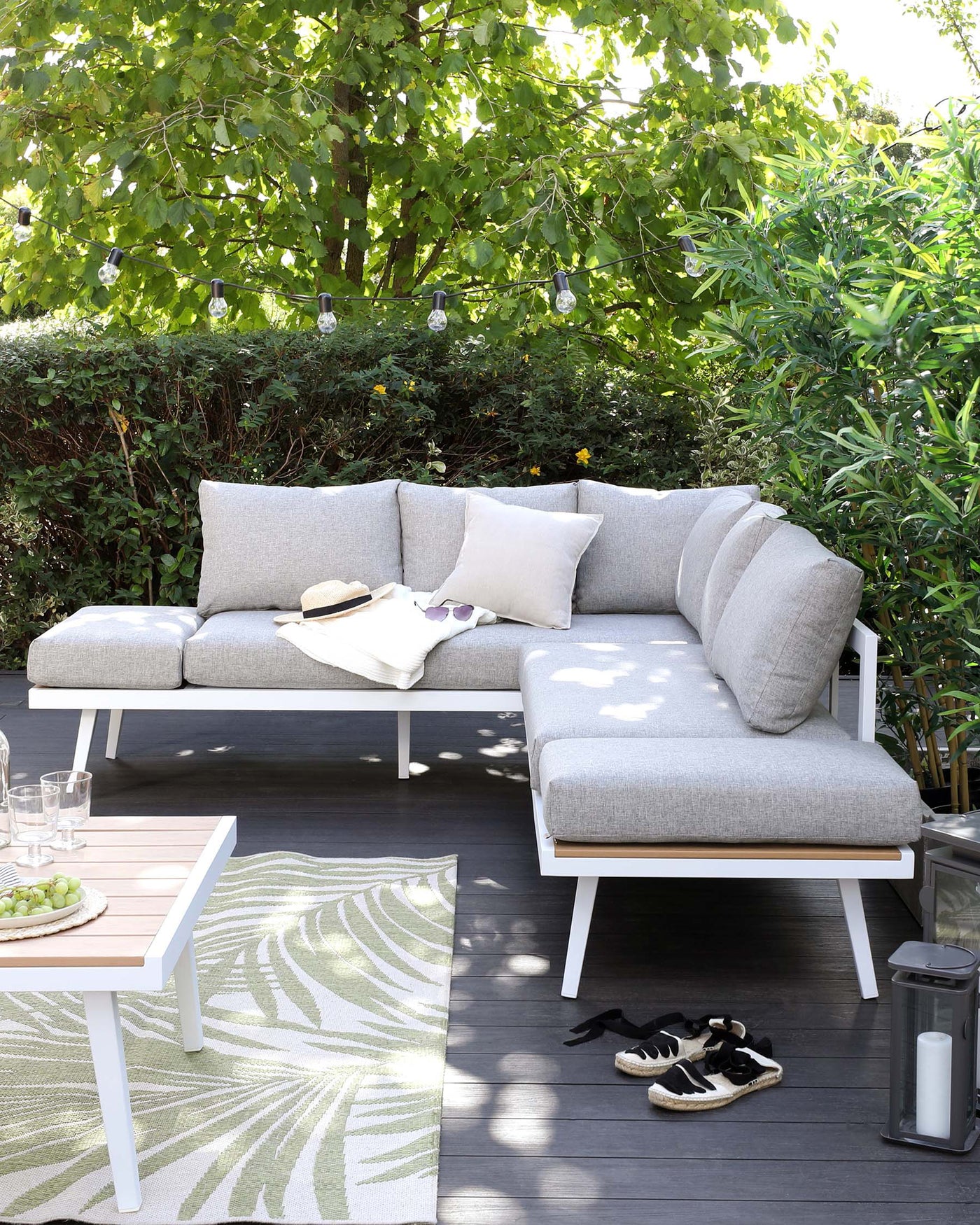 Outdoor furniture set on a wooden deck with a light grey upholstered corner sofa and a white minimalist coffee table, complemented by patterned area rug and ambient string lights above.
