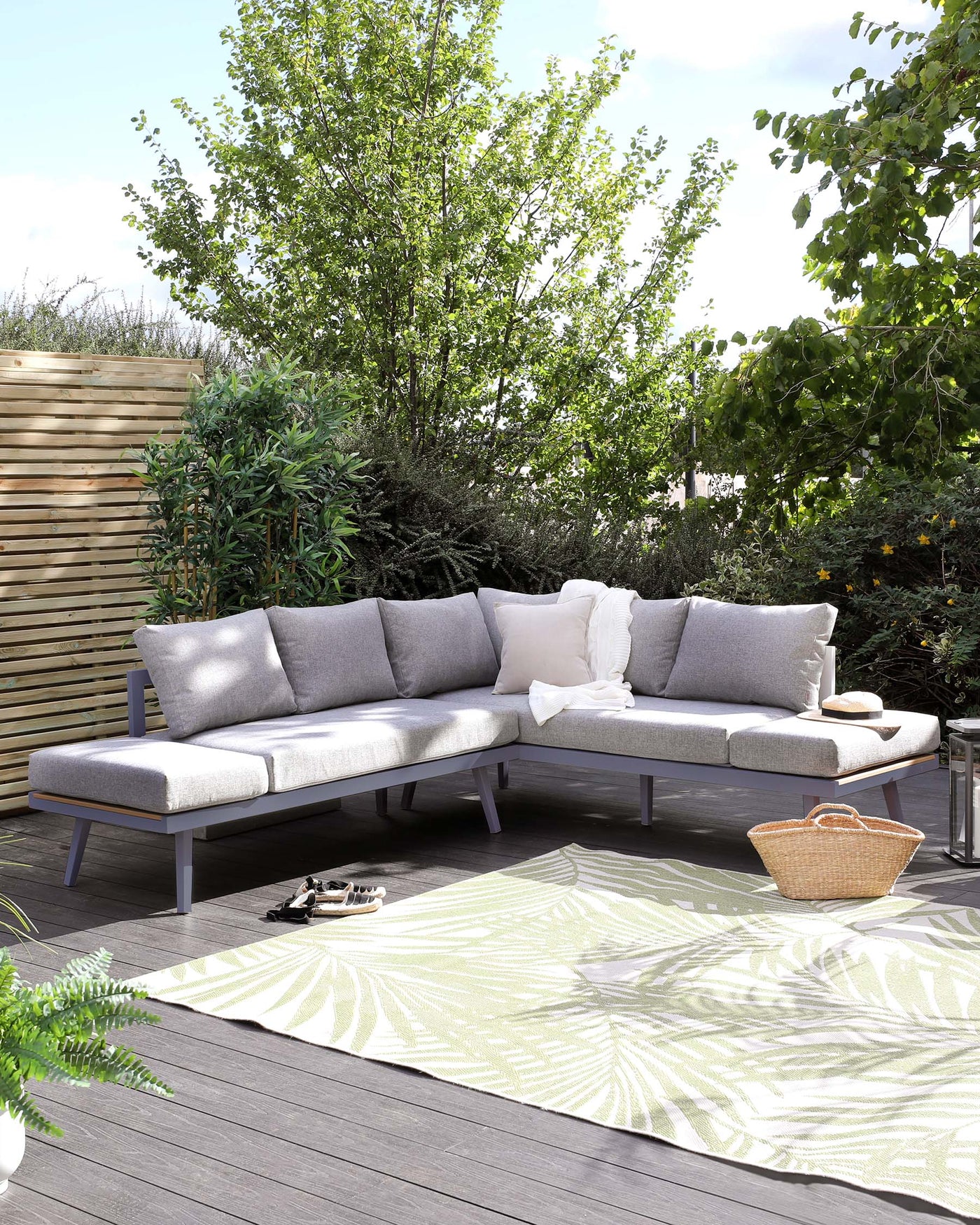 Modern outdoor L-shaped sectional sofa with light grey cushions, a chaise lounge on the right side, and a metal frame in a dark finish set on a wooden deck with decorative outdoor pillows, accompanied by a small metal side table. A patterned light green and white area rug lies beneath the arrangement, enhancing the outdoor setting.