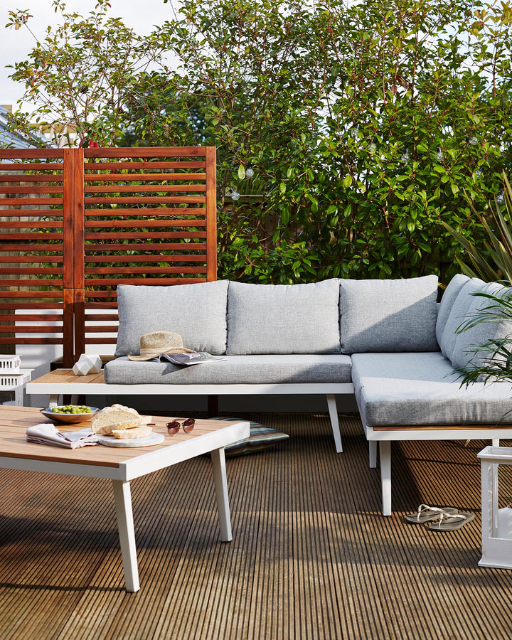 Modern outdoor furniture set featuring a sectional sofa with light grey cushions, a white metal frame, and teak wood accents. A matching low white coffee table with a teak wood top sits in front, adorned with a casual spread of food and a magazine, creating an inviting terrace or patio setting.