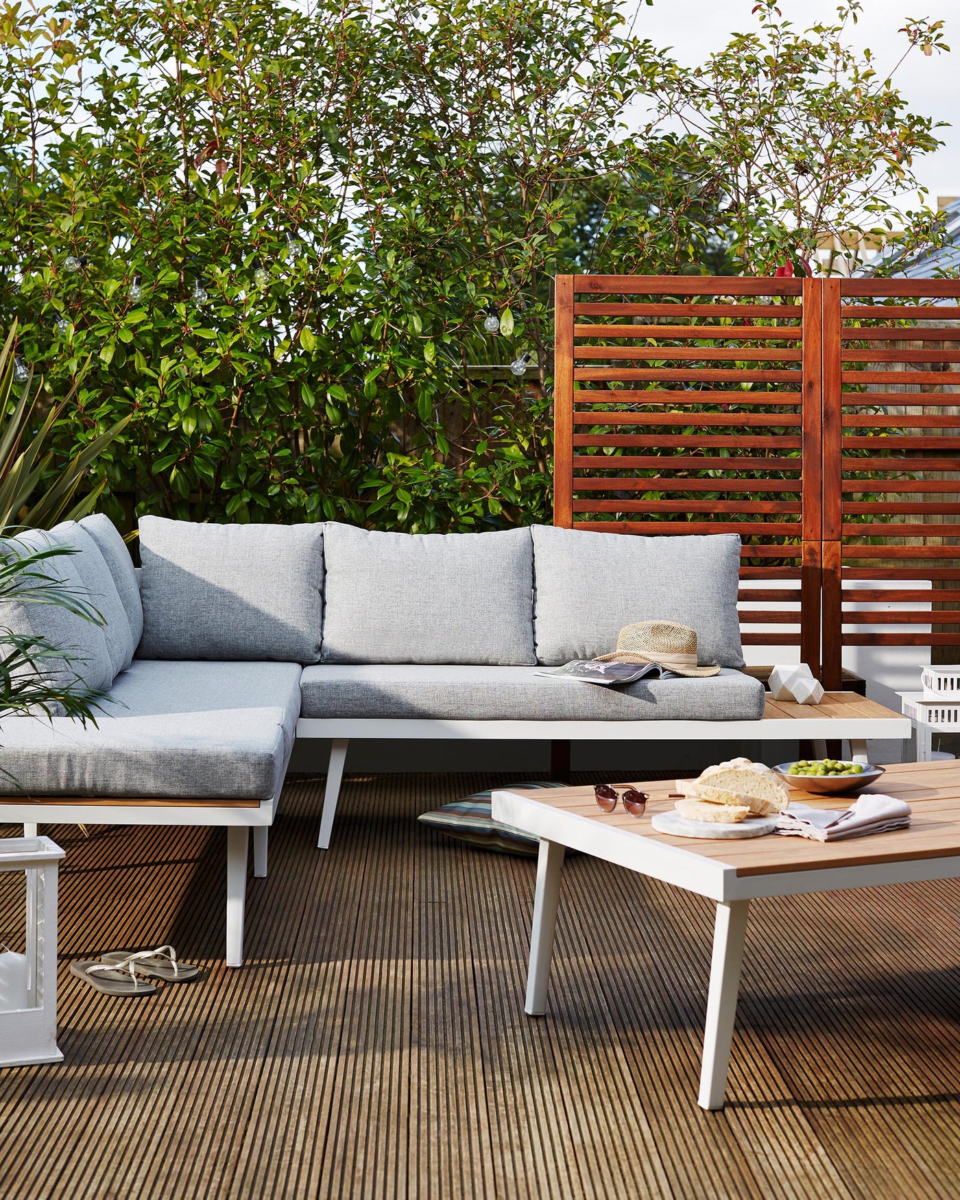 Modern outdoor furniture set featuring a sectional sofa with grey cushions and a white frame, complemented by a two-toned wood and white coffee table. A small white side table is also present, alongside decorative items such as pillows, books, and a straw hat. The set is arranged on a wooden deck with lush greenery in the background, providing a cosy and inviting outdoor living space.