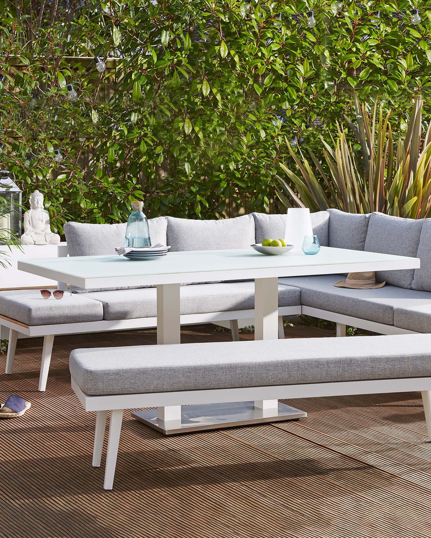 Outdoor contemporary furniture set featuring a white rectangular dining table with a sleek modern design, surrounded by a matching white corner sectional sofa and two benches, all with grey cushions. The set is arranged on a brown textured outdoor rug, complemented by decorative elements like a buddha statue, blue glassware, and a bowl of limes, creating an inviting al fresco dining space.