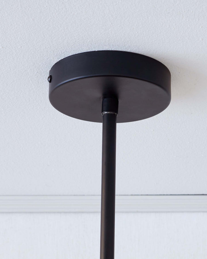 The image depicts the ceiling mount and stem of a pendant light, showing a black circular canopy attached to the ceiling from which a slender black rod extends, presumably leading to a light fixture out of frame. This image does not display furniture.