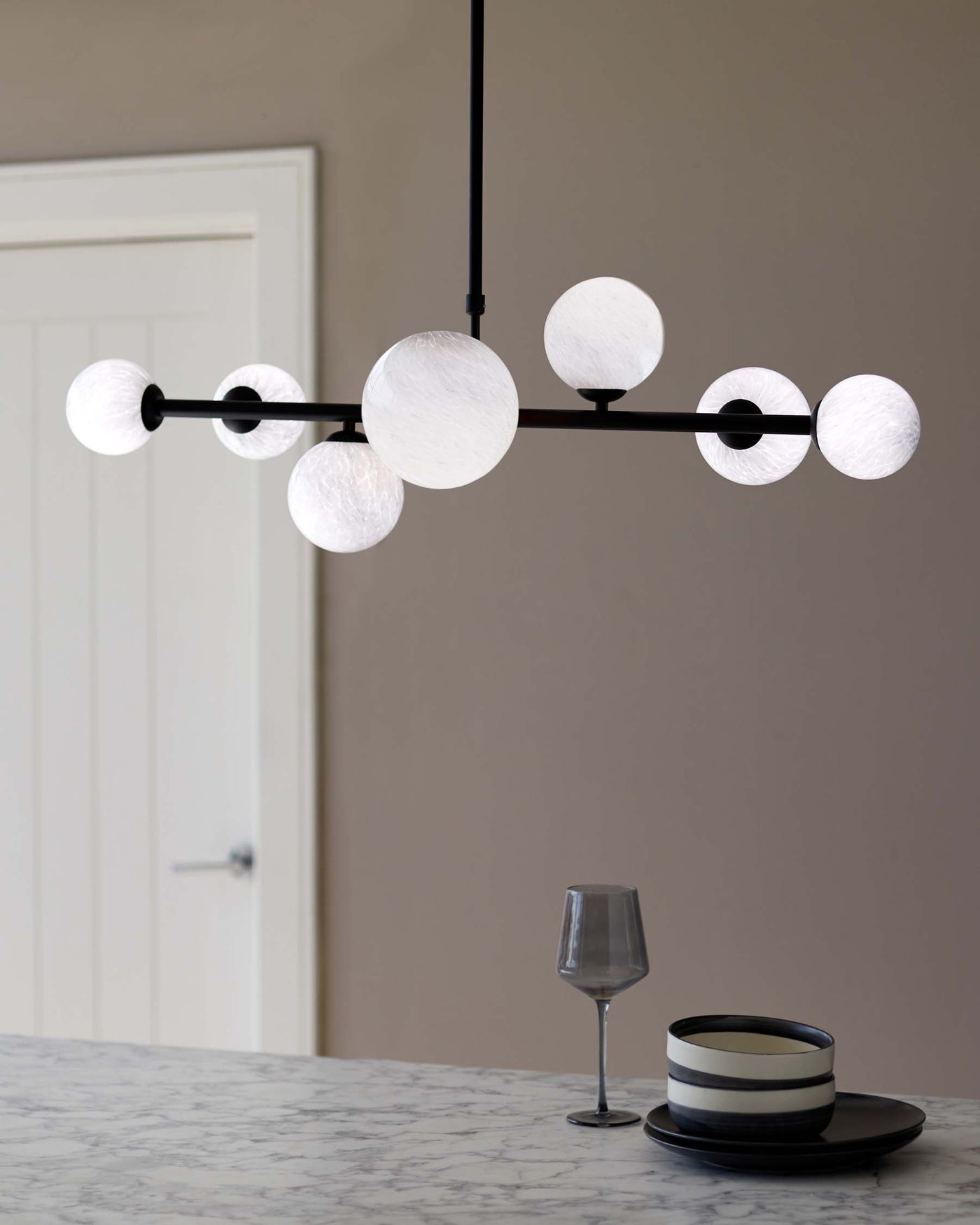 A minimalist marble tabletop with a set of two dark, contrasting-rimmed plates and a smoke grey wine glass displayed under an elegant, modern chandelier featuring a linear black fixture with evenly spaced opaque white globes.