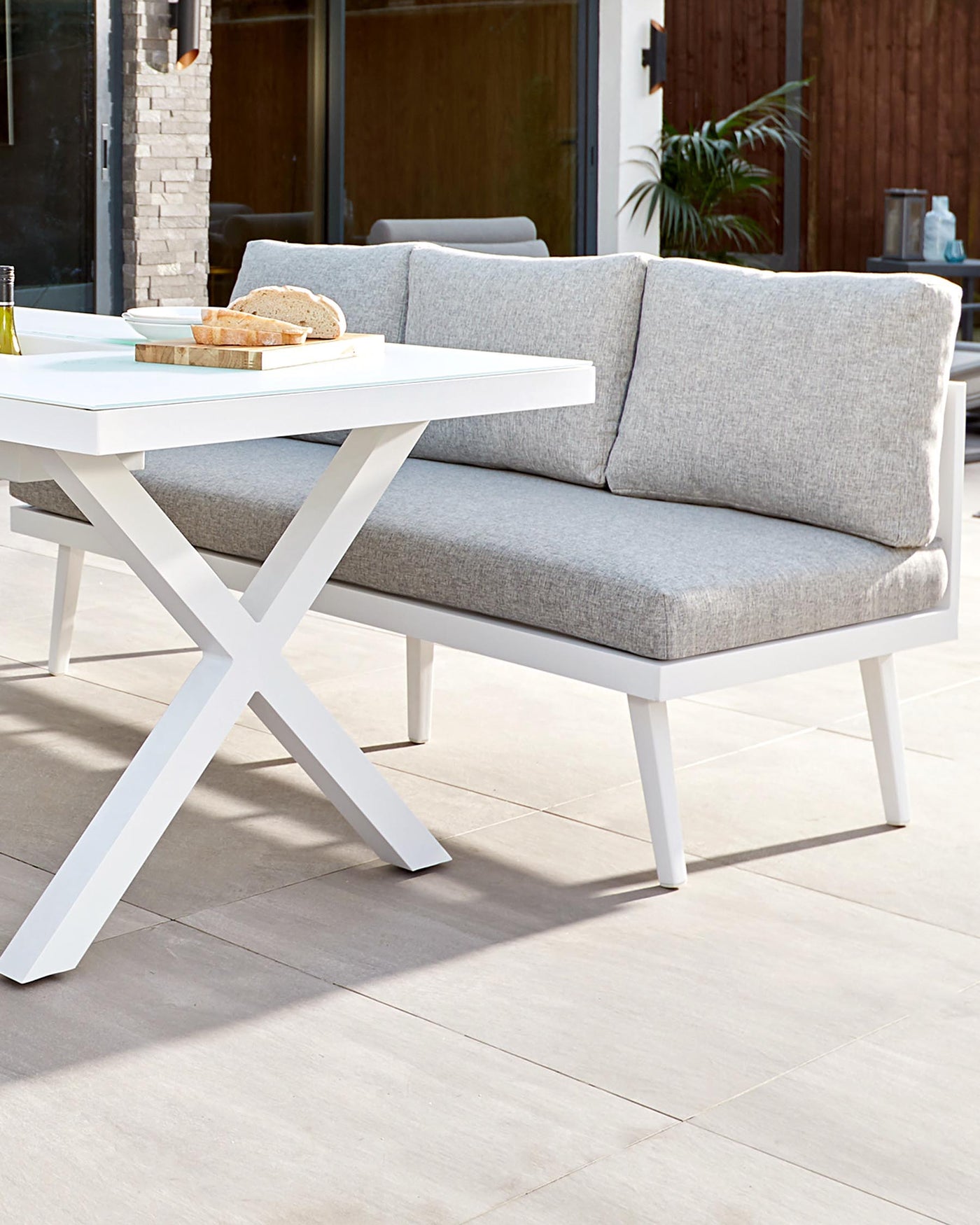 Modern outdoor furniture featuring a white rectangular coffee table with a unique X-shaped base, paired with a grey cushioned bench with a sleek white frame. The setting suggests a comfortable and stylish patio arrangement.