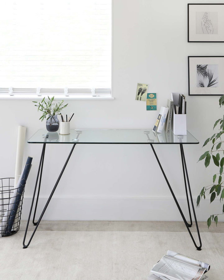 A minimalist modern desk with a clear glass tabletop and sleek black metal hairpin legs.