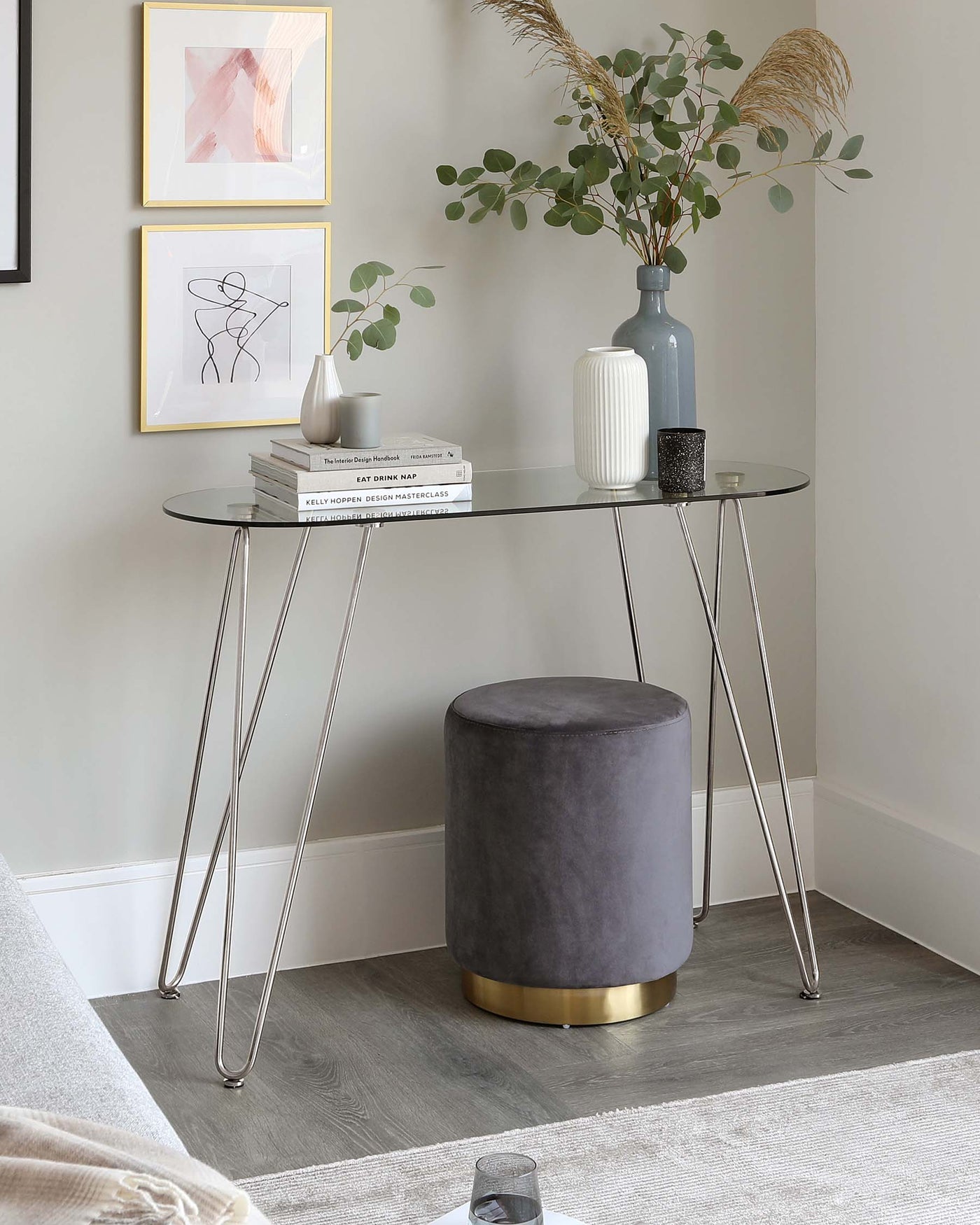 An elegant, modern console table with a sleek, round glass top and slender, tapered metallic legs alongside a chic, grey upholstered ottoman with a golden base.